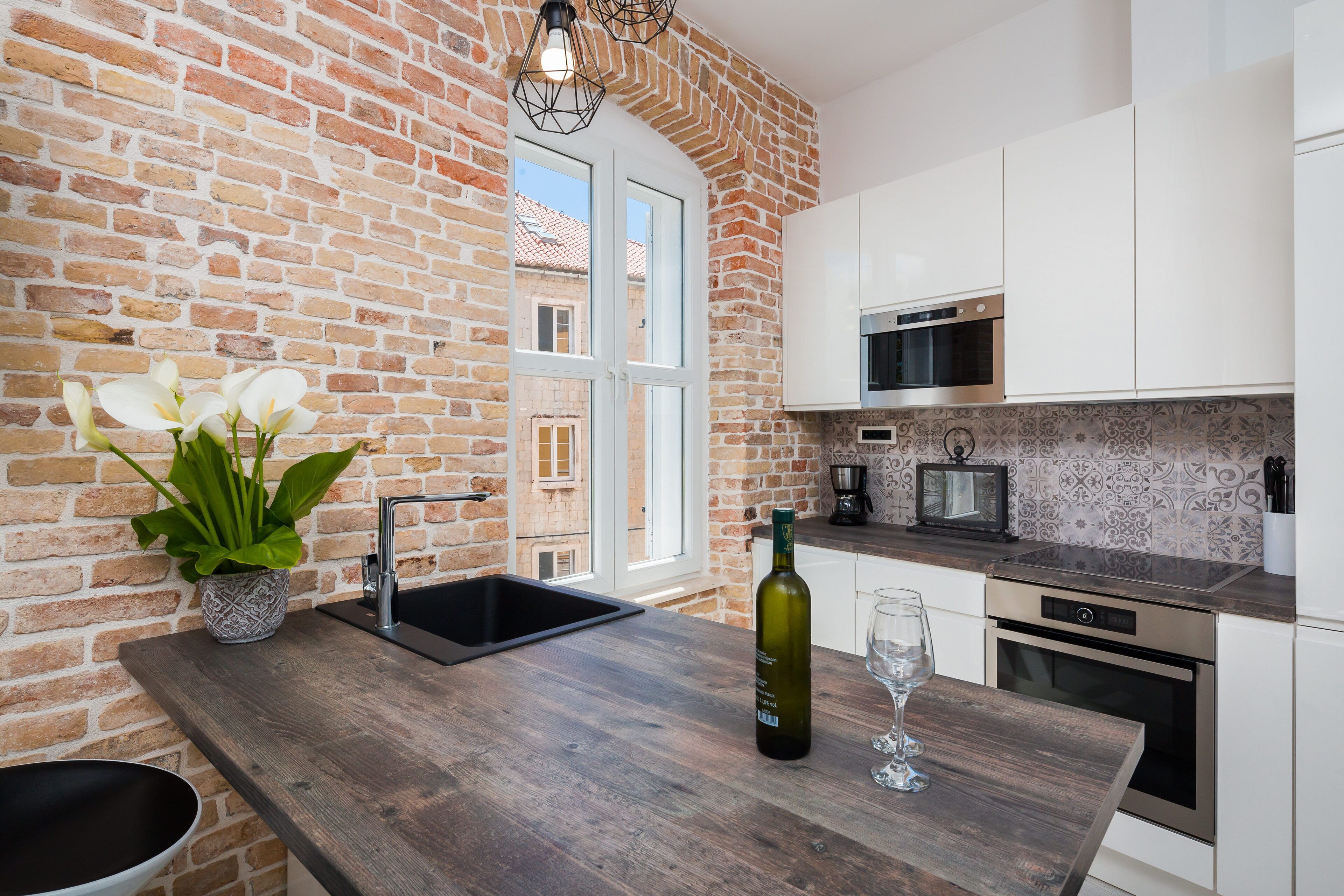 Une cuisine avec un mur en briques, des armoires blanches, un plan de travail en bois, un évier, une bouteille de vin et un verre.