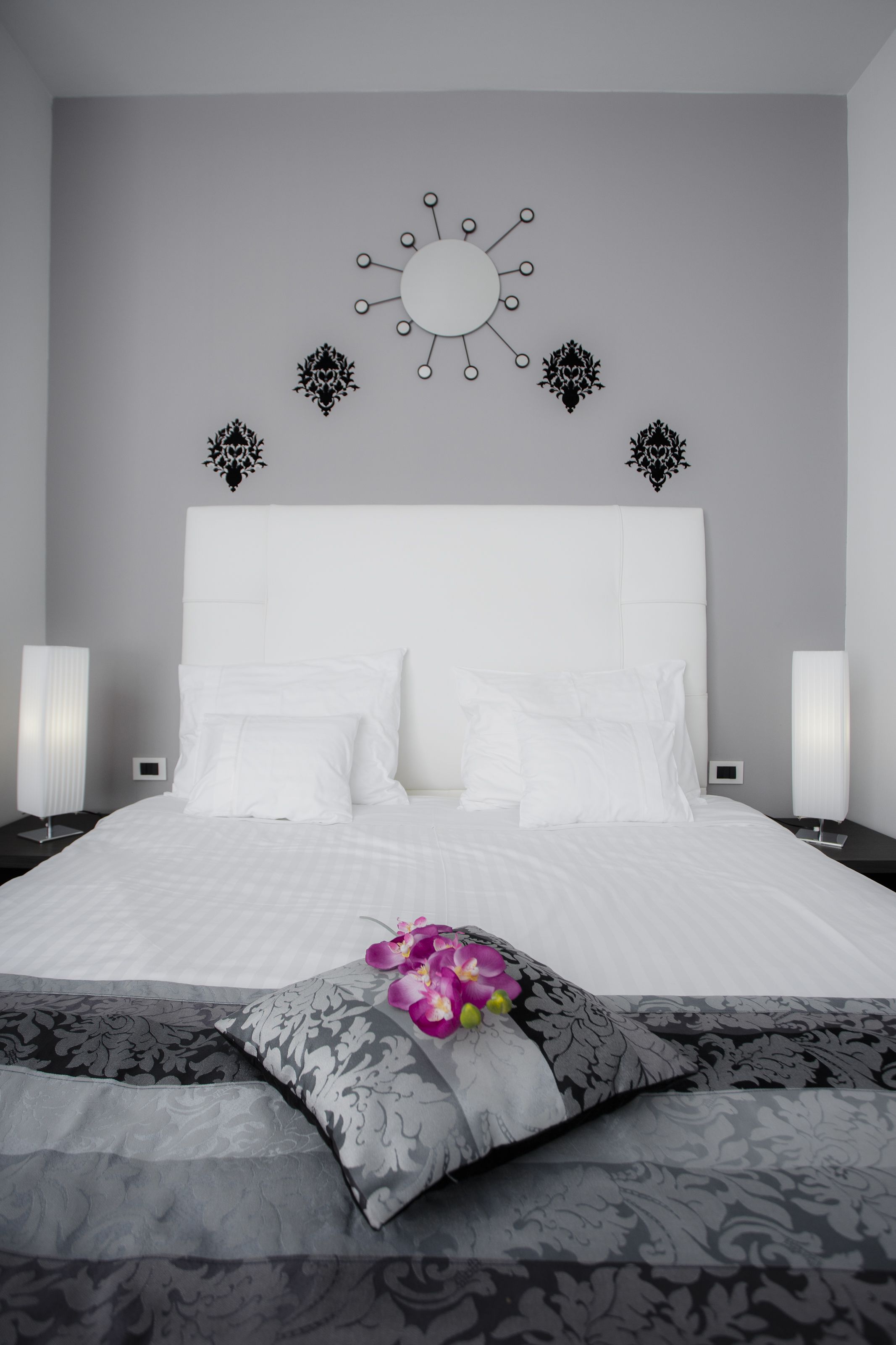 A modern bedroom with a large bed, white bedding, decorative wall art, and a single purple orchid.