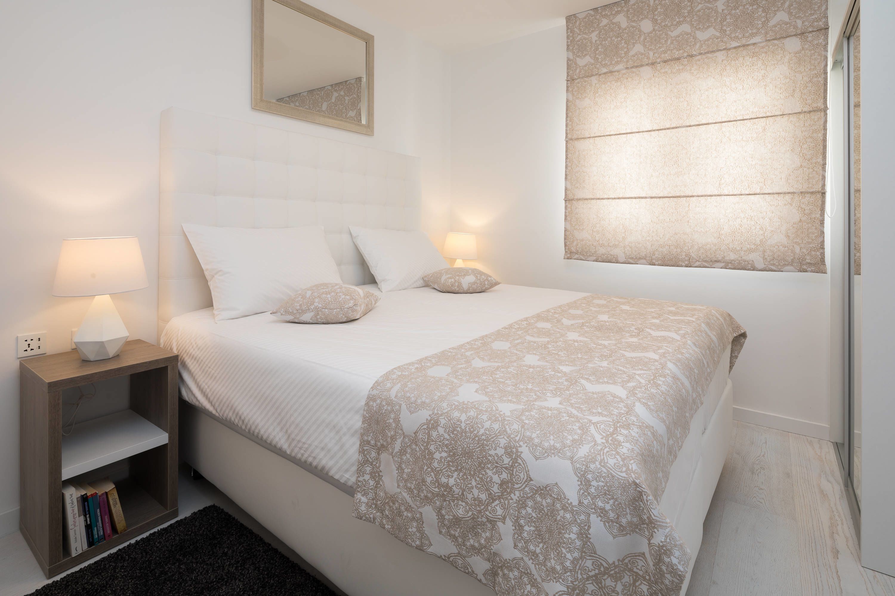 A bedroom with a bed, bedside lamps, a nightstand with books, a mirror, and a window with blinds.