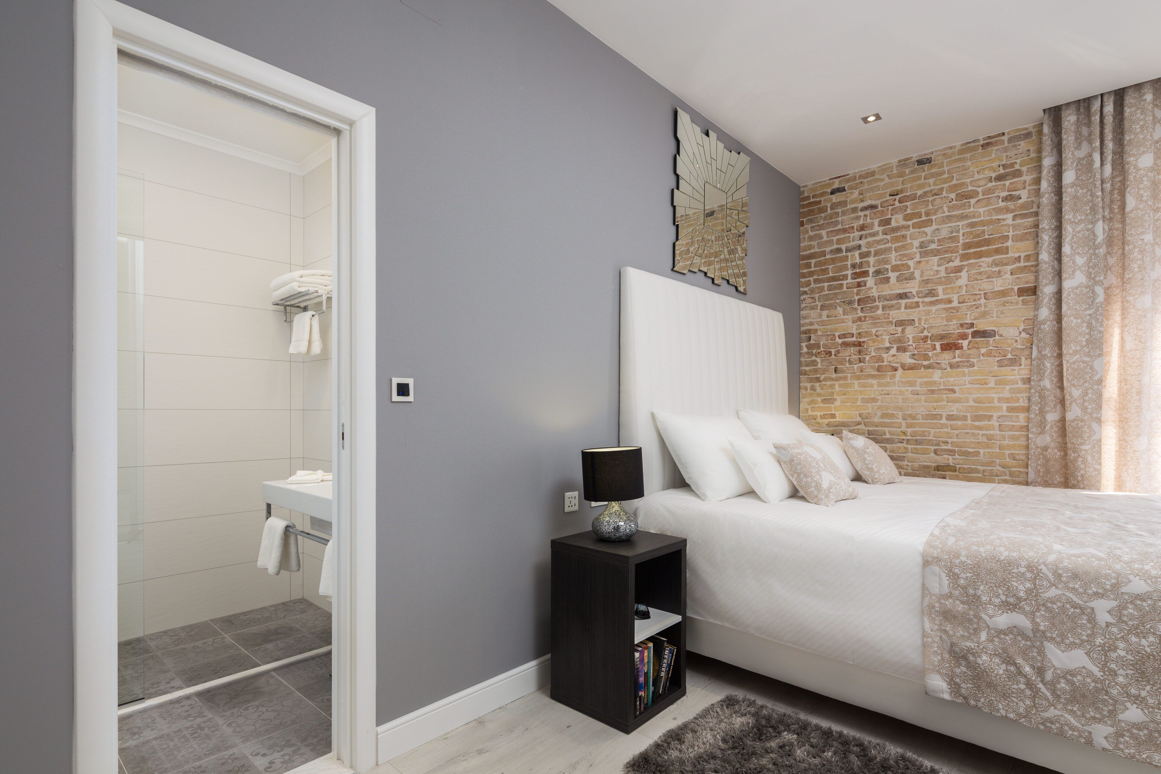 A bedroom with a bed, a nightstand, a mirror, and a brick wall.