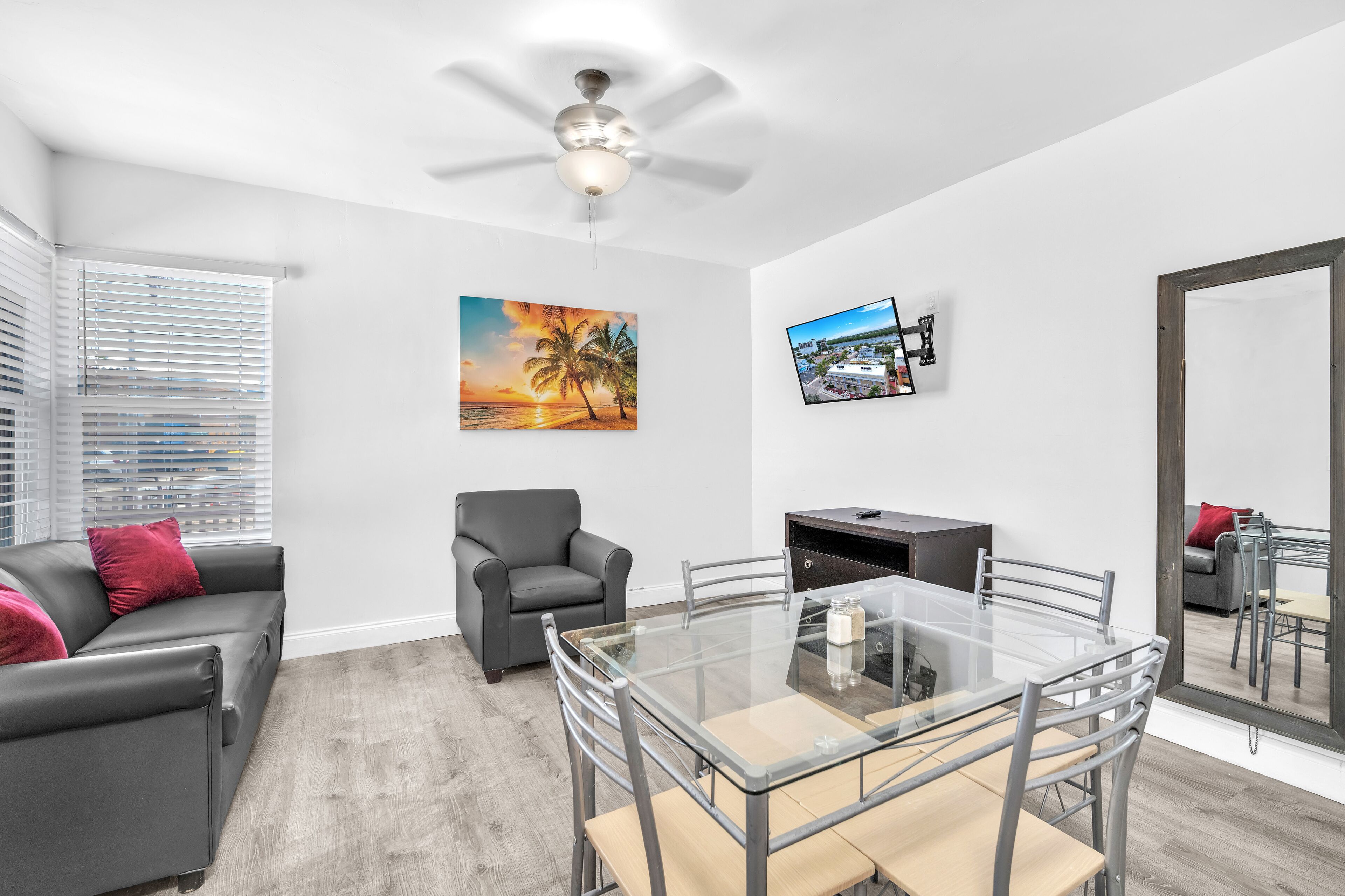 A modern living room with a glass dining table, grey sofa, and a ceiling fan.