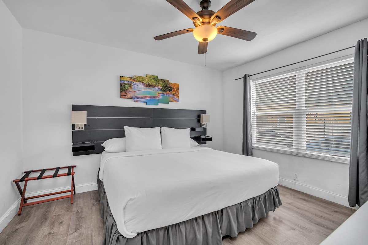 Un dormitorio moderno con una cama grande, un ventilador de techo, un cuadro en la pared y una ventana con persianas.