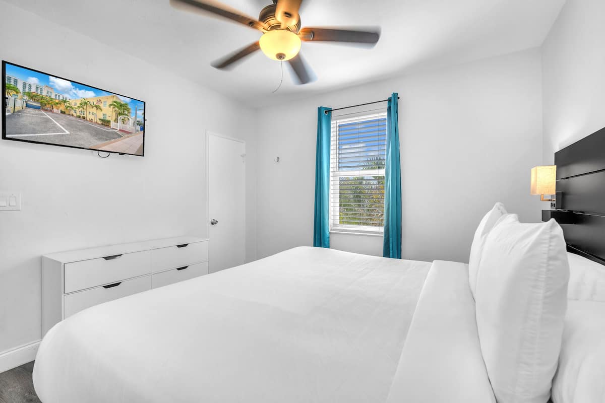 Un dormitorio con cama, ventilador de techo, televisor en la pared, ventana con cortinas y lámpara de noche.