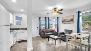 A modern living room with a gray sofa, a coffee table, and a dining table with chairs. There is a ceiling fan, a window with blue curtains, and a painting on the wall. - Laguna Blu Beach (Hollywood)