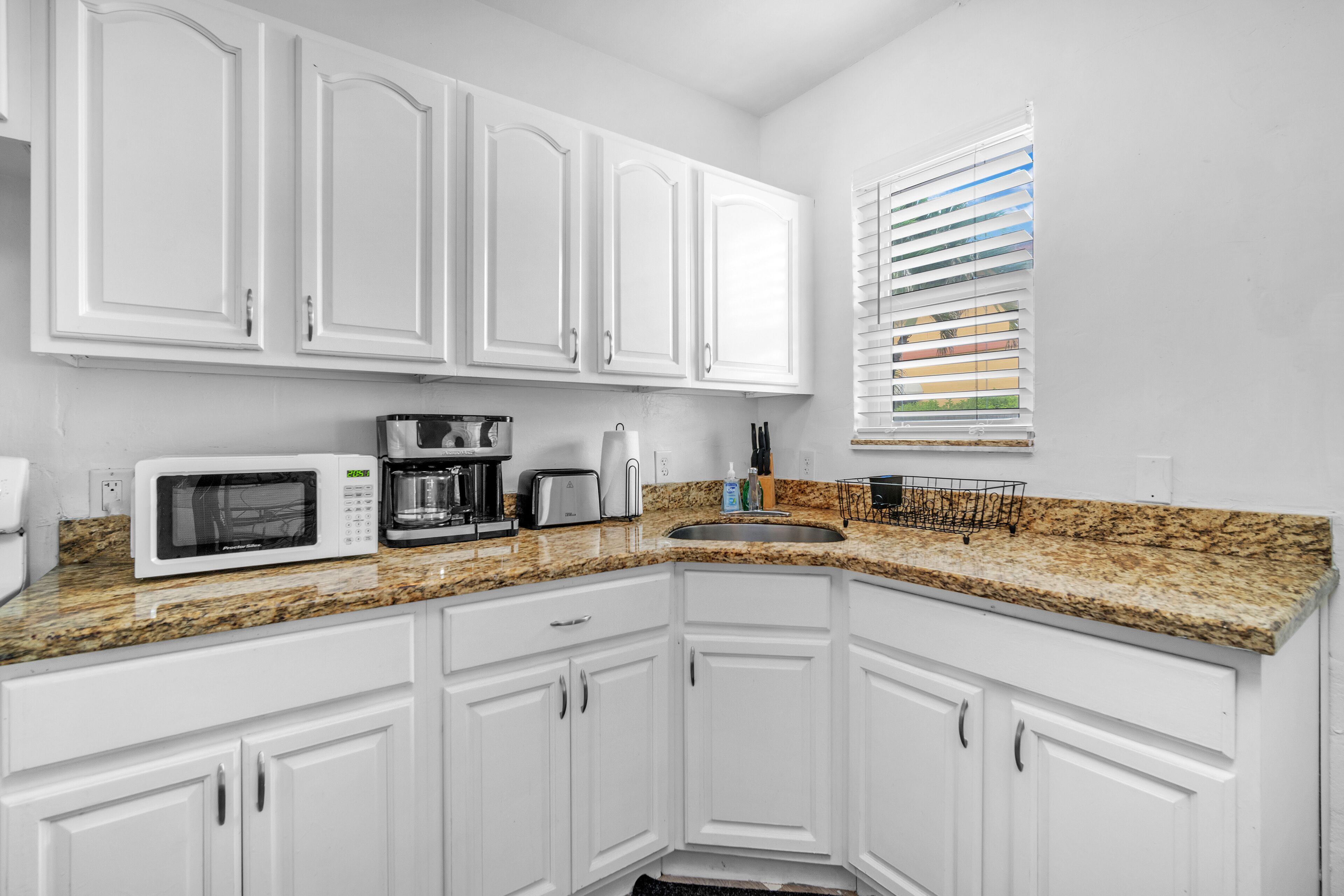 A modern kitchen with white cabinets, a granite countertop, a microwave, a coffee maker, and a window with blinds.