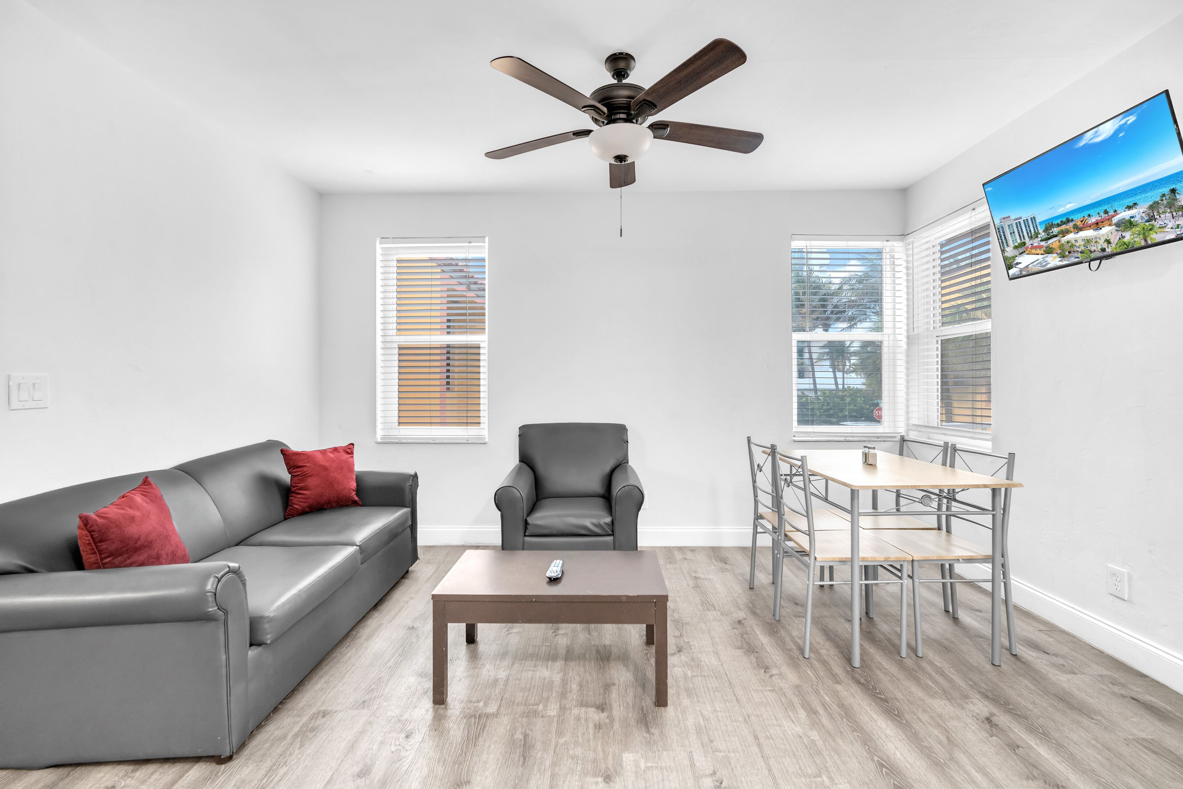 A living room with a gray sofa, a gray armchair, a wooden coffee table, a dining table with chairs, a ceiling fan, and a TV on the wall.
