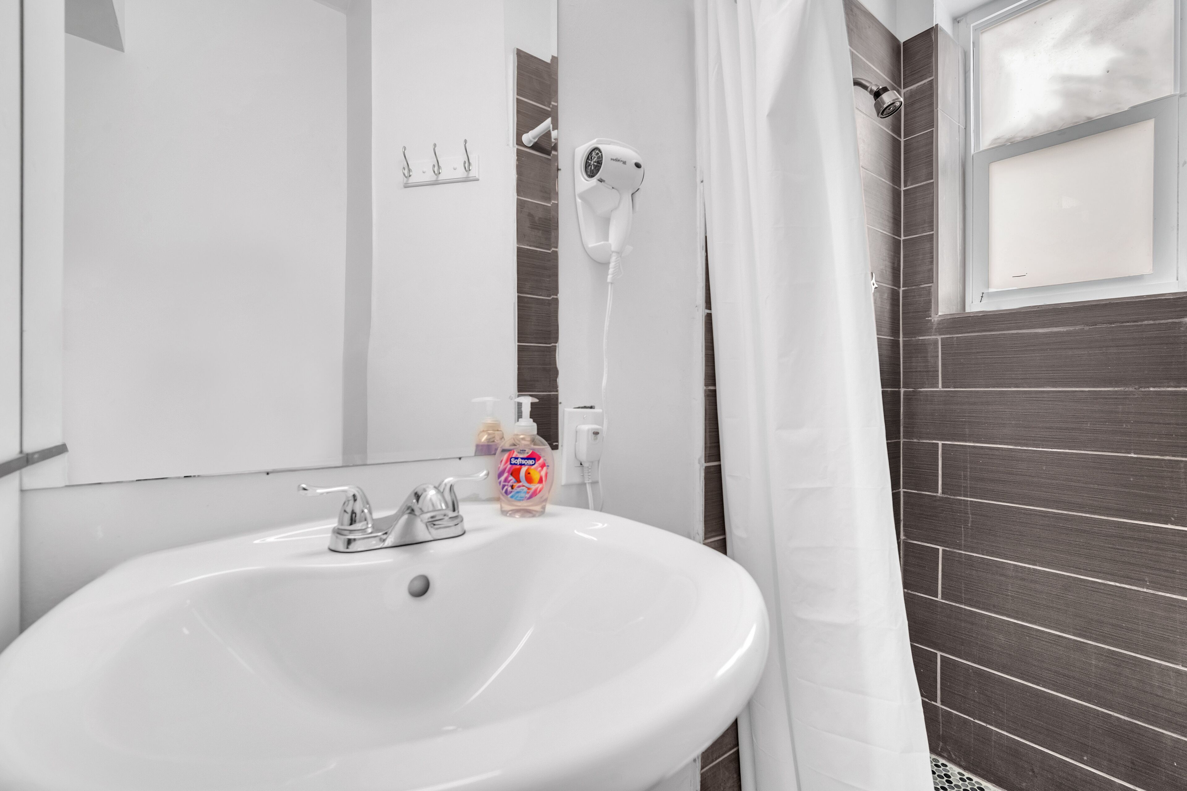 Une salle de bain avec un lavabo blanc, un sèche-cheveux fixé au mur et une fenêtre avec un rideau.