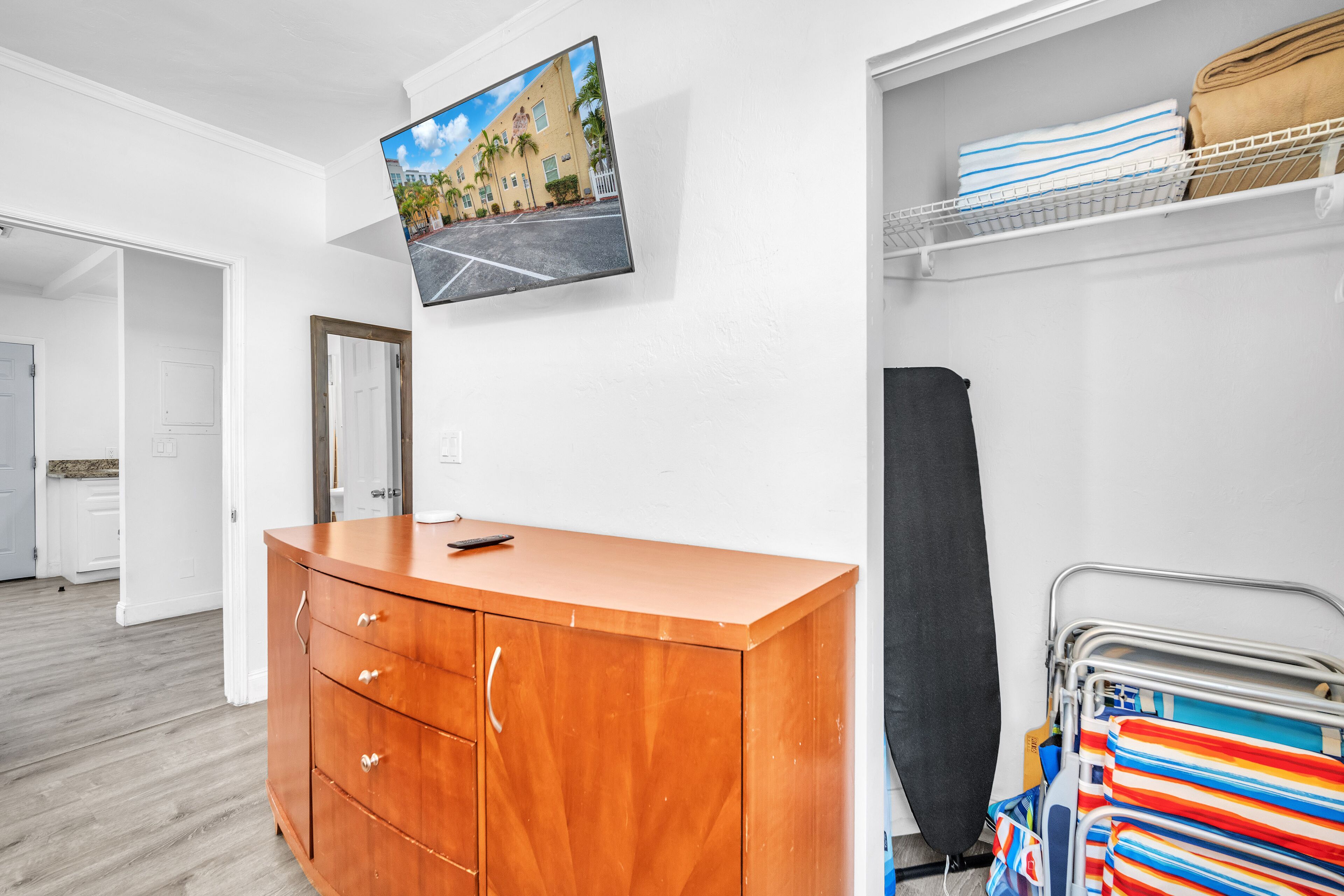 A hotel room with a wooden dresser, a flat-screen TV mounted on the wall, a metal rack with towels, and a door leading to another room.