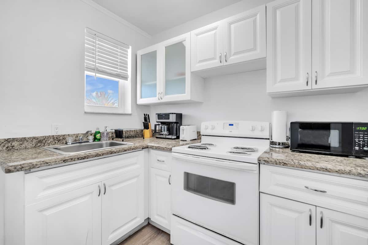 Una cocina moderna con armarios blancos, encimera de granito, estufa, microondas y ventana con persianas.