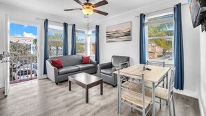 A spacious living room with a gray sofa, a wooden coffee table, and a dining table with metal chairs. A ceiling fan is visible, and there are large windows with blue curtains. - Laguna Blu Beach (Hollywood)