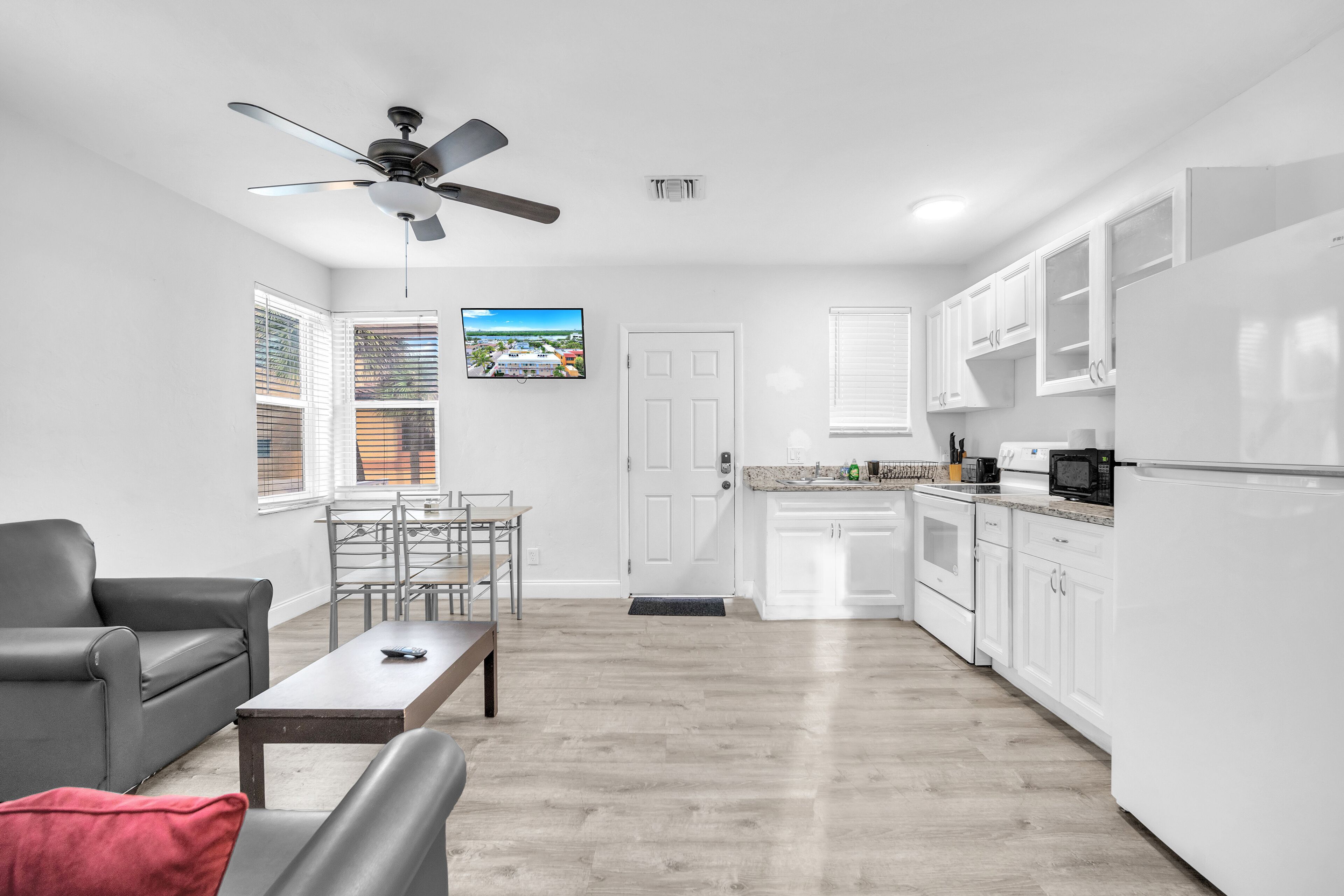 A modern kitchen with white cabinets, a built-in microwave, and a refrigerator. There is a dining area with a table and chairs, and a ceiling fan above.