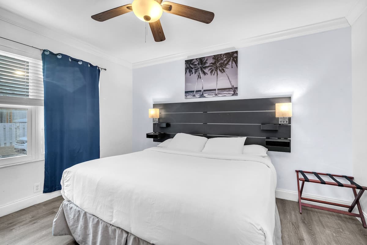 Un dormitorio con cama, ventilador de techo, mesita de noche con lámpara, ventana con persianas y un cuadro colgado en la pared.