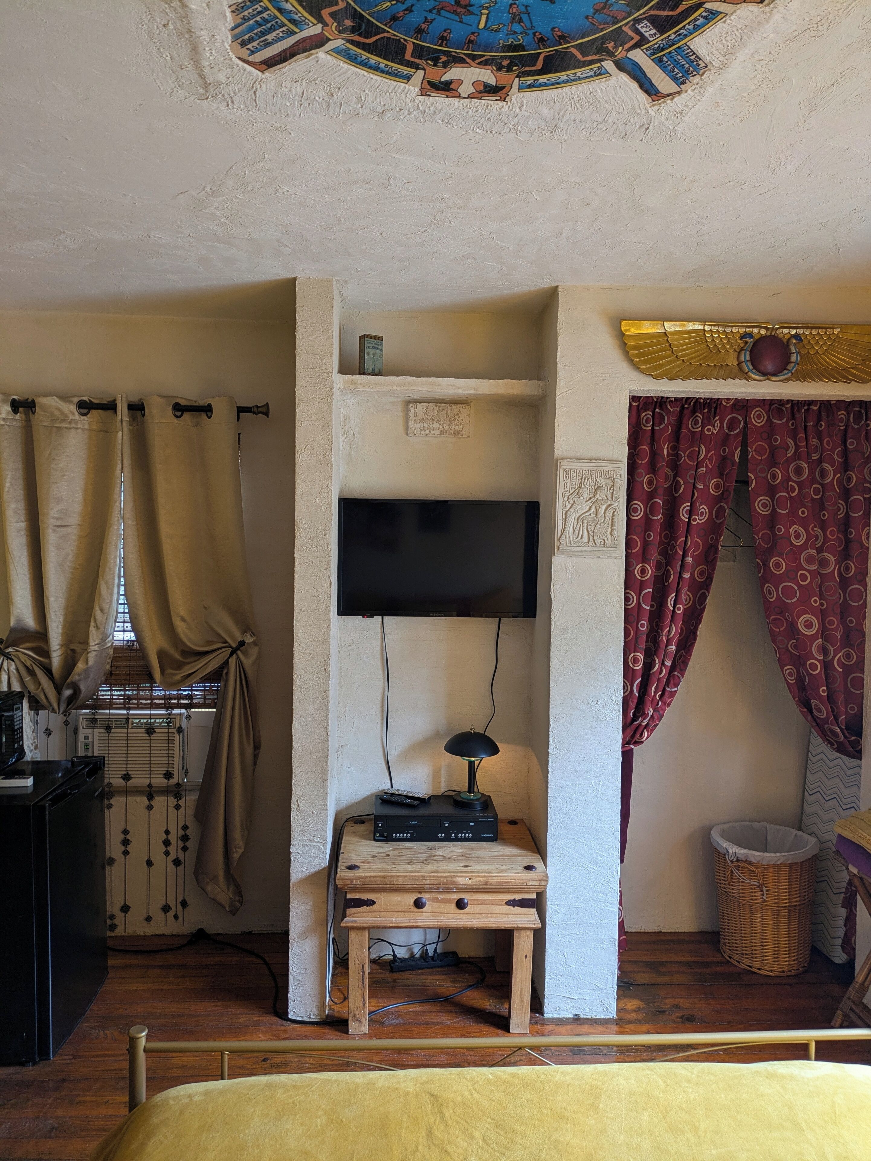 Una habitación con televisión montada en la pared, una pequeña mesa de madera con una lámpara, una cama con cubrecama amarillo y un techo decorativo.
