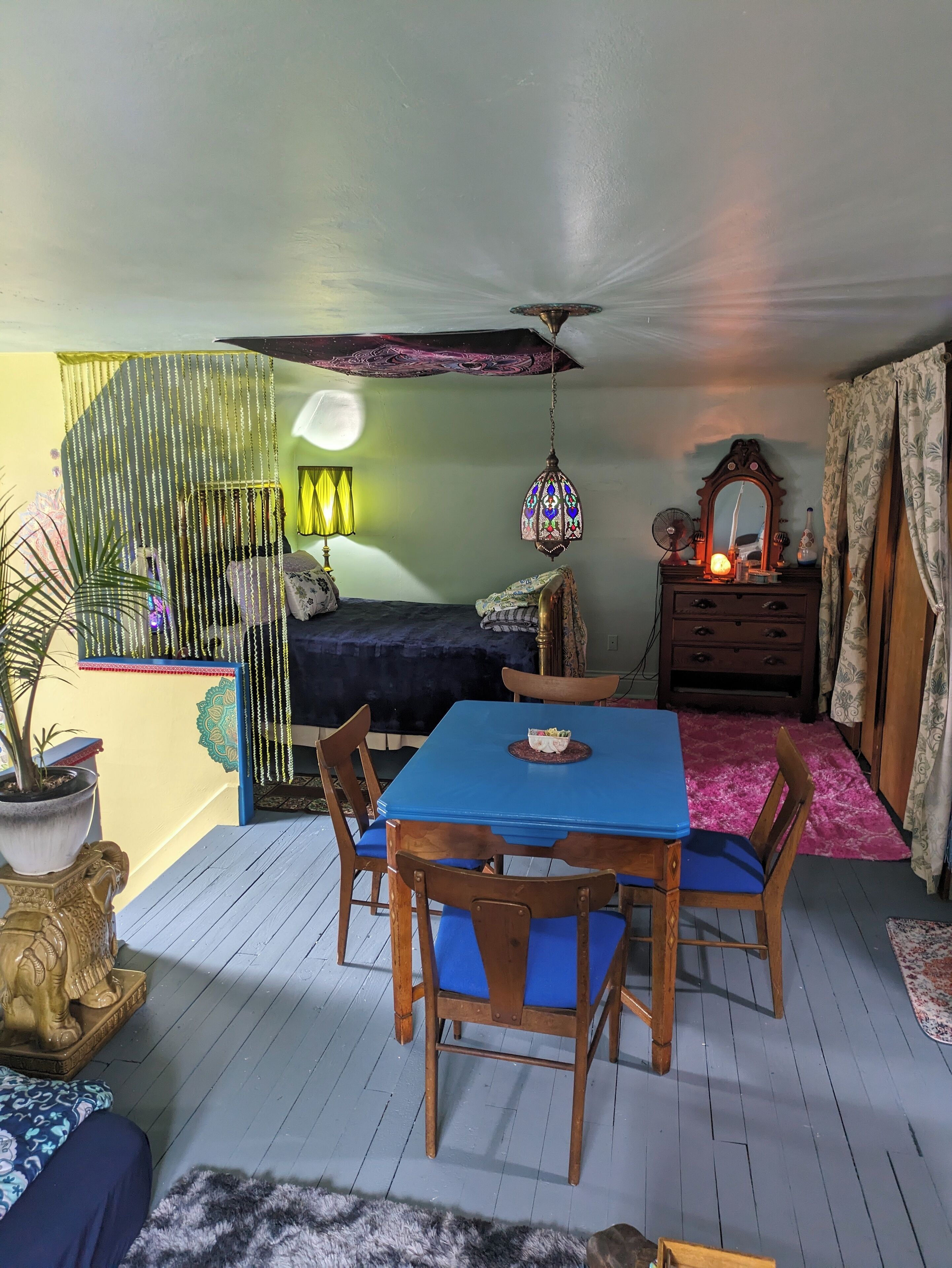 A room with a dining table, chairs, a bed, a dresser, and a ceiling fan.
