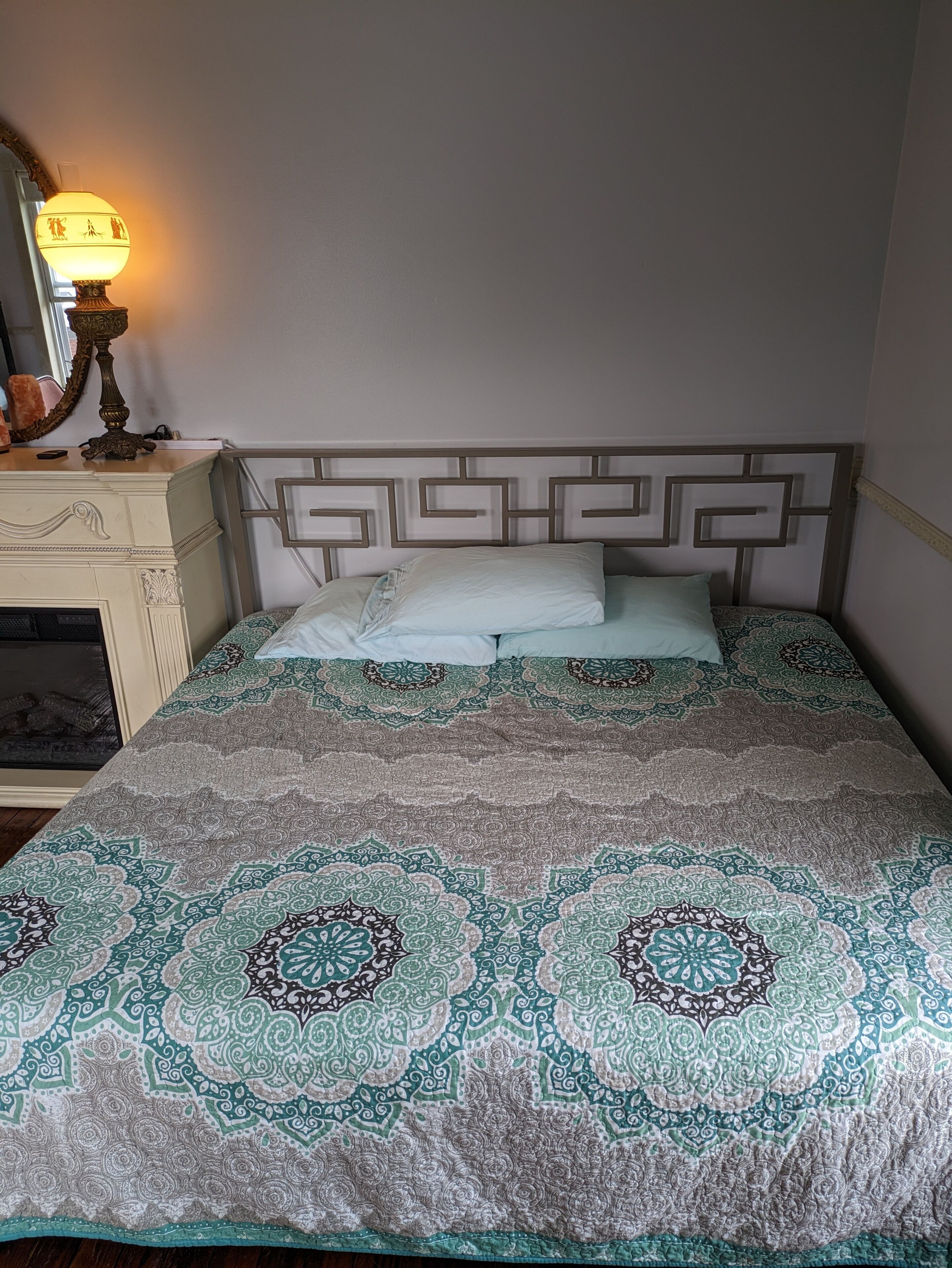A bed with a patterned bedspread, a bedside table with a lamp, and a mirror on the wall.