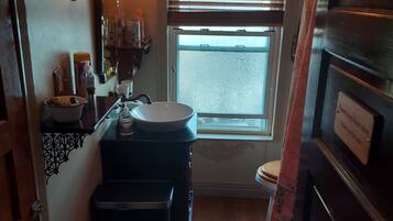 A compact bathroom with a sink, vanity, and toilet. There is a window with blinds, a mirror with wall sconces, and a door with a sign.