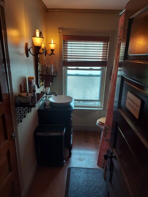 Un baño compacto con lavamanos, tocador e inodoro. Hay una ventana con persianas, un espejo con apliques de pared, y una puerta con un cartel.