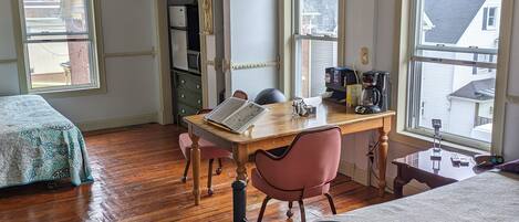 Une pièce dotée d’un plancher en bois, d’un lit avec un cadre en métal, d’un bureau avec une chaise et de deux fenêtres ornées de moulures décoratives.