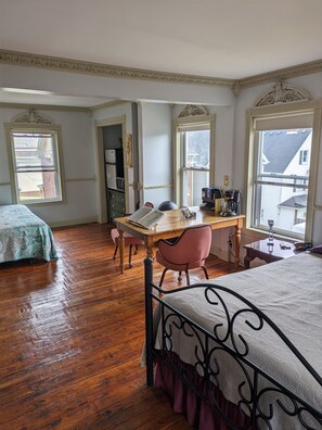 Una habitación con piso de madera, una cama con estructura metálica, un escritorio con silla y dos ventanas con molduras decorativas.