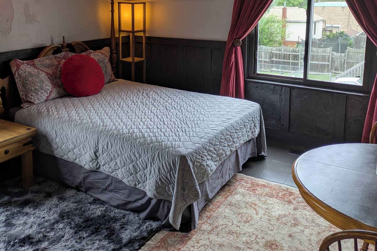 A bedroom with a bed, a bedside table, a lamp, a window with curtains, a painting, and a view of the outside.