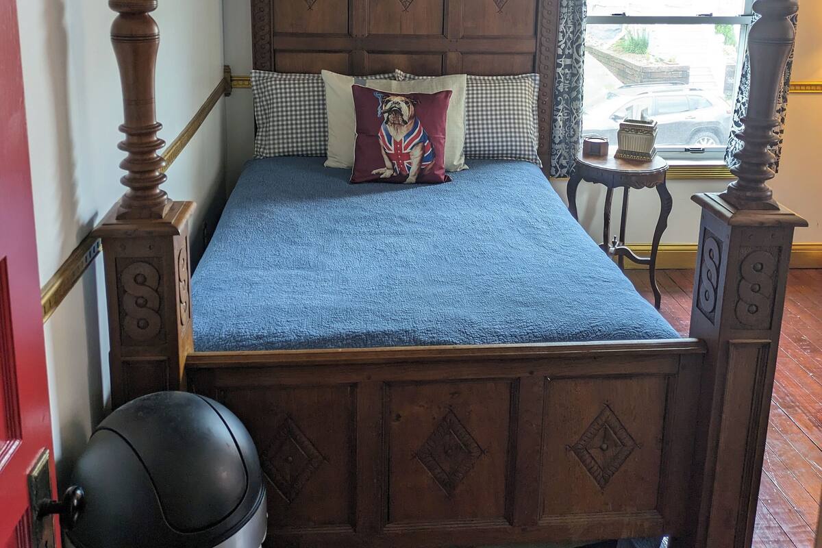 A four-poster bed with a checkered headboard, a blue bedspread, and a pillow with a Union Jack design.