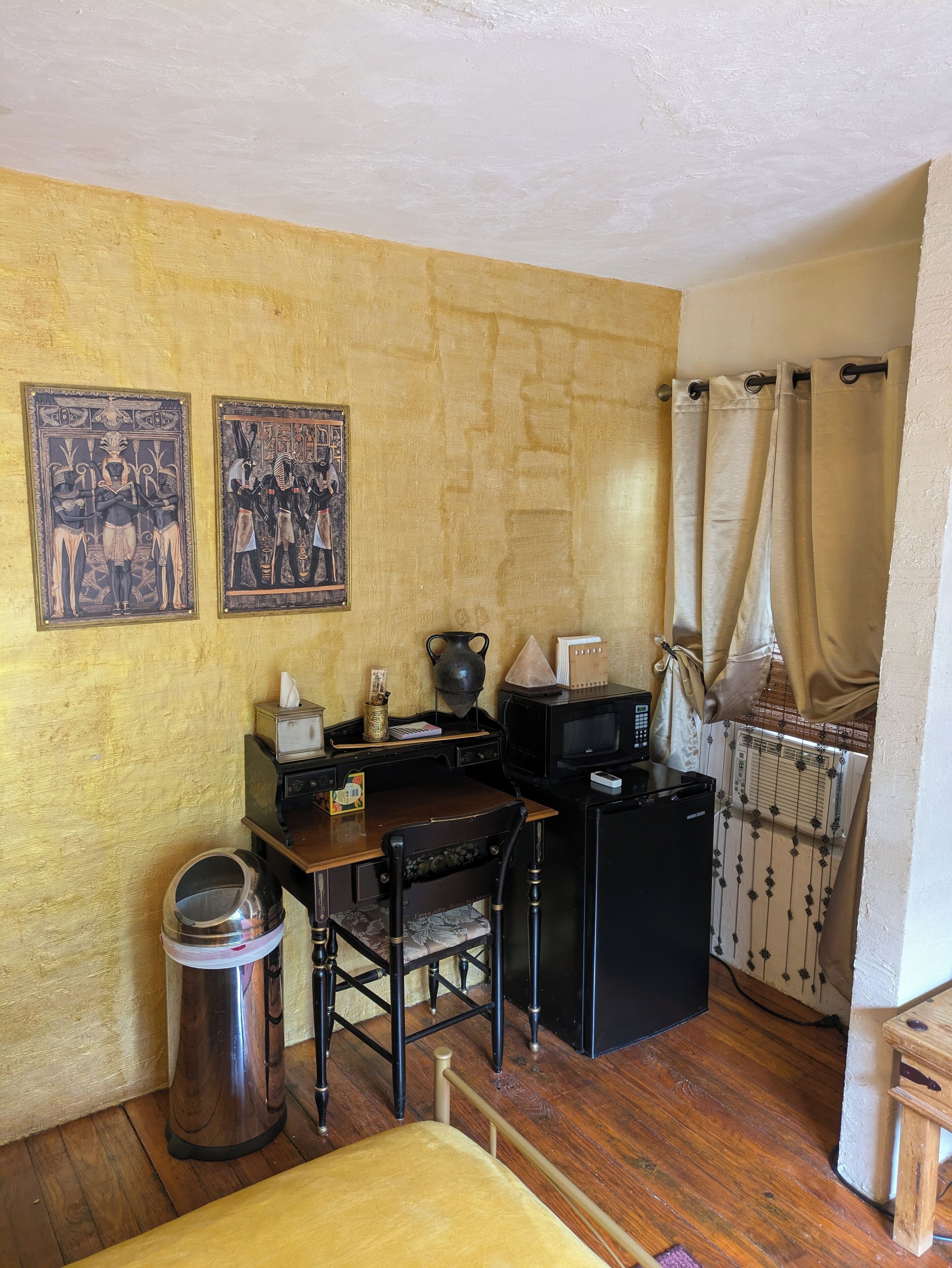 Cozinha compacta com mesa de madeira, geladeira preta e micro-ondas. Há duas imagens penduradas na parede retratando cenas do antigo Egito.