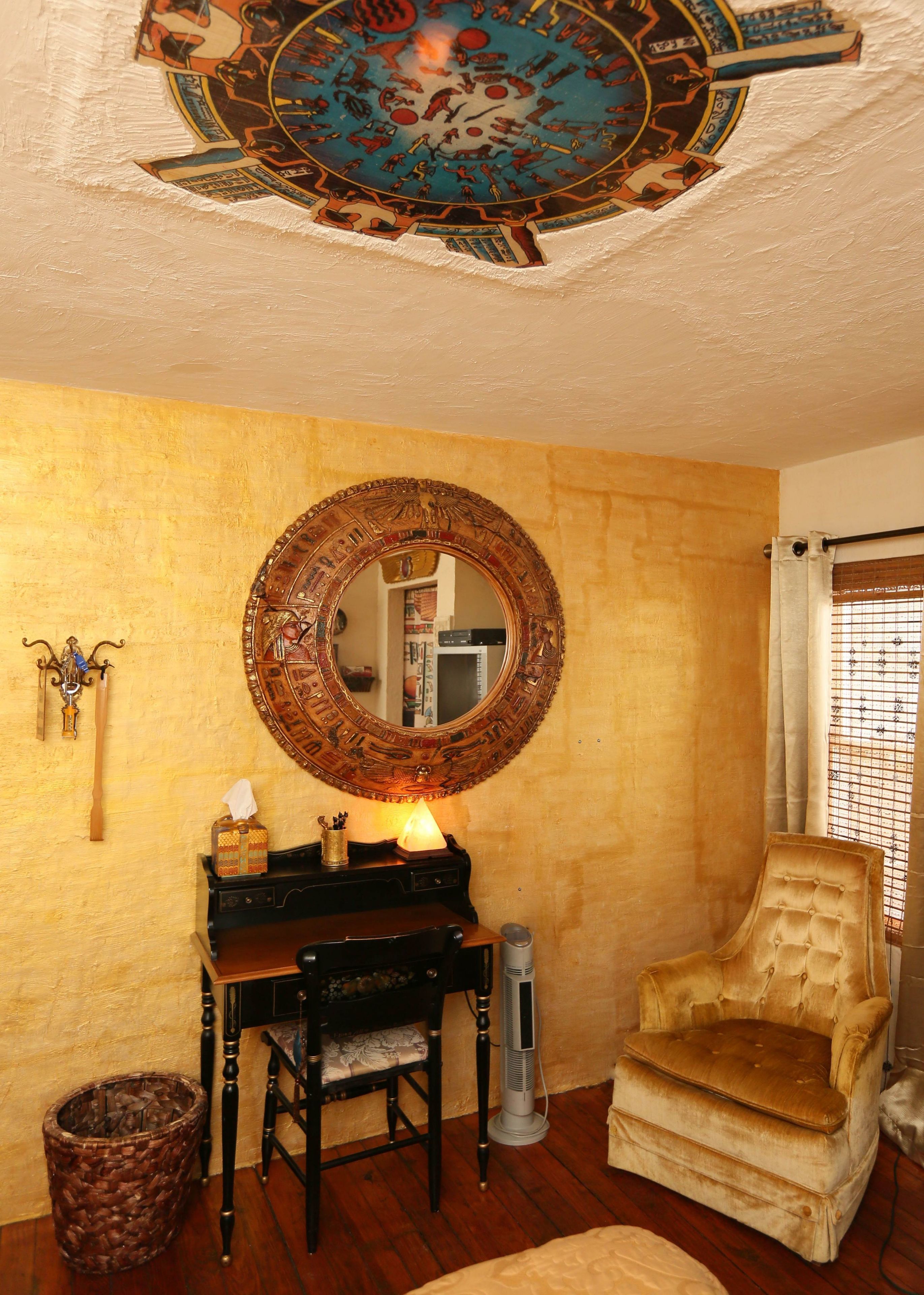 A room with a round mirror, a wooden desk, a cushioned armchair, a standing heater, and a woven basket.
