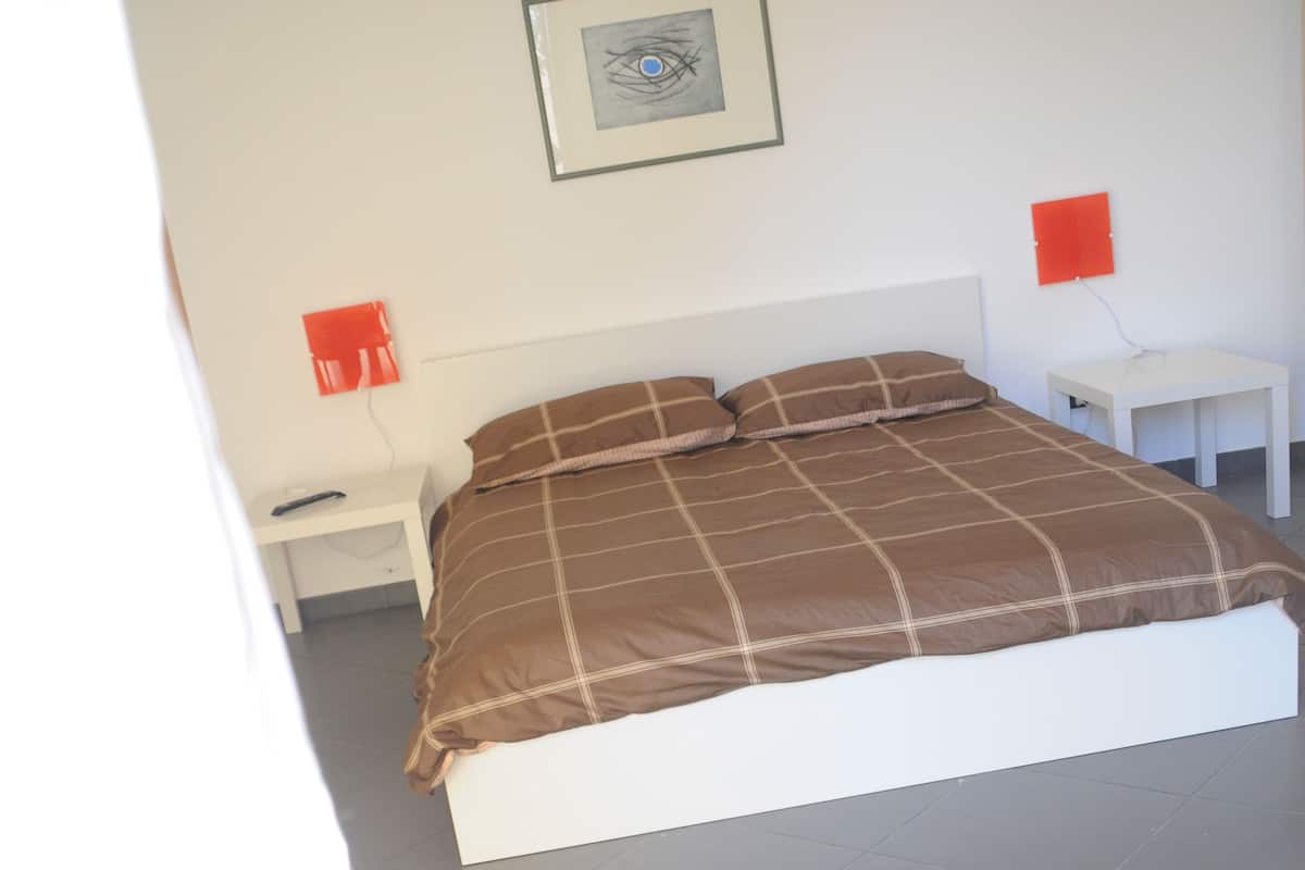 A bedroom with a bed, a nightstand, a bedside table, and a wall-mounted light fixture.