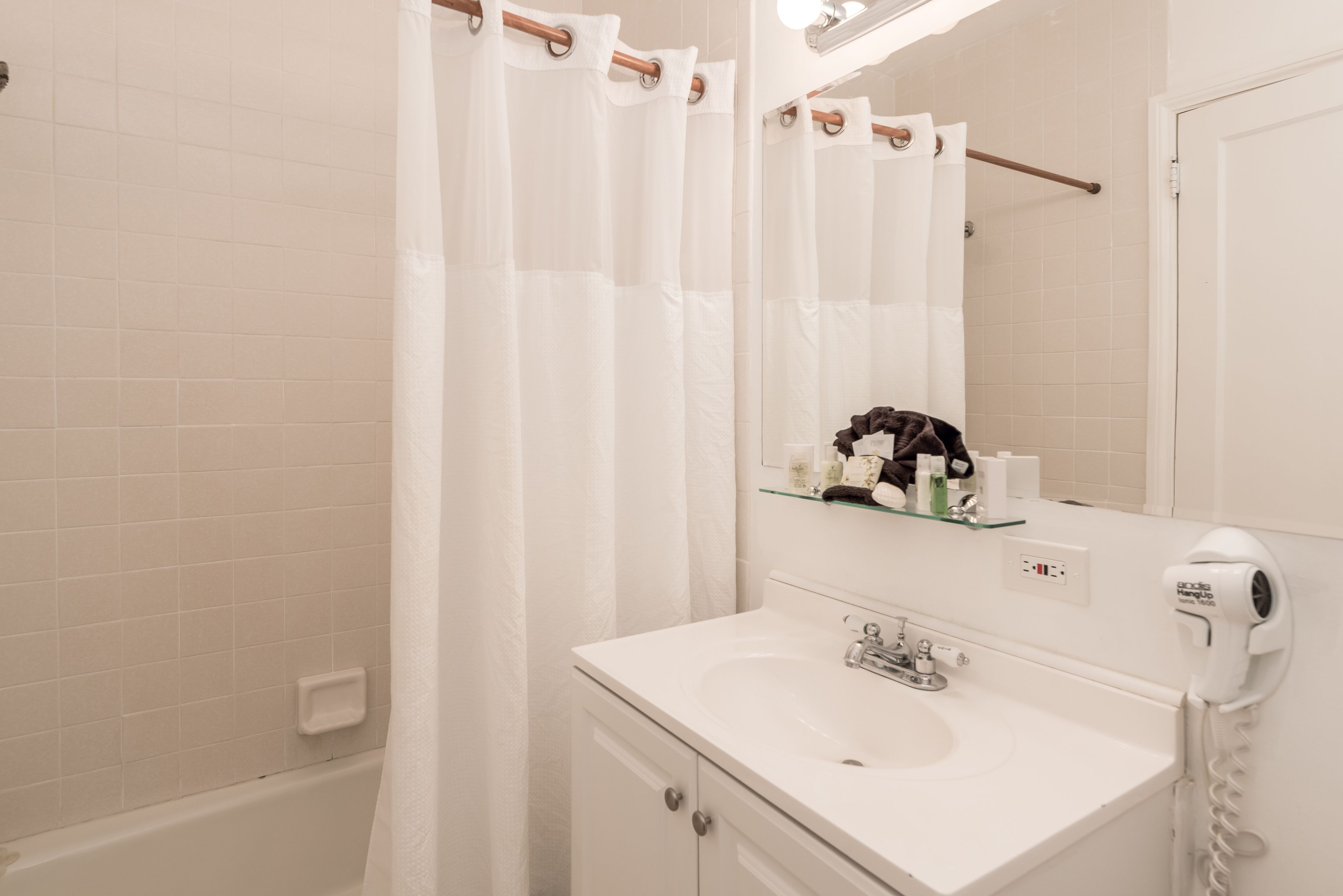 Un baño moderno con un lavamanos blanco, un estante de vidrio con artículos de aseo, una cortina de ducha y un secador de pelo en la pared.