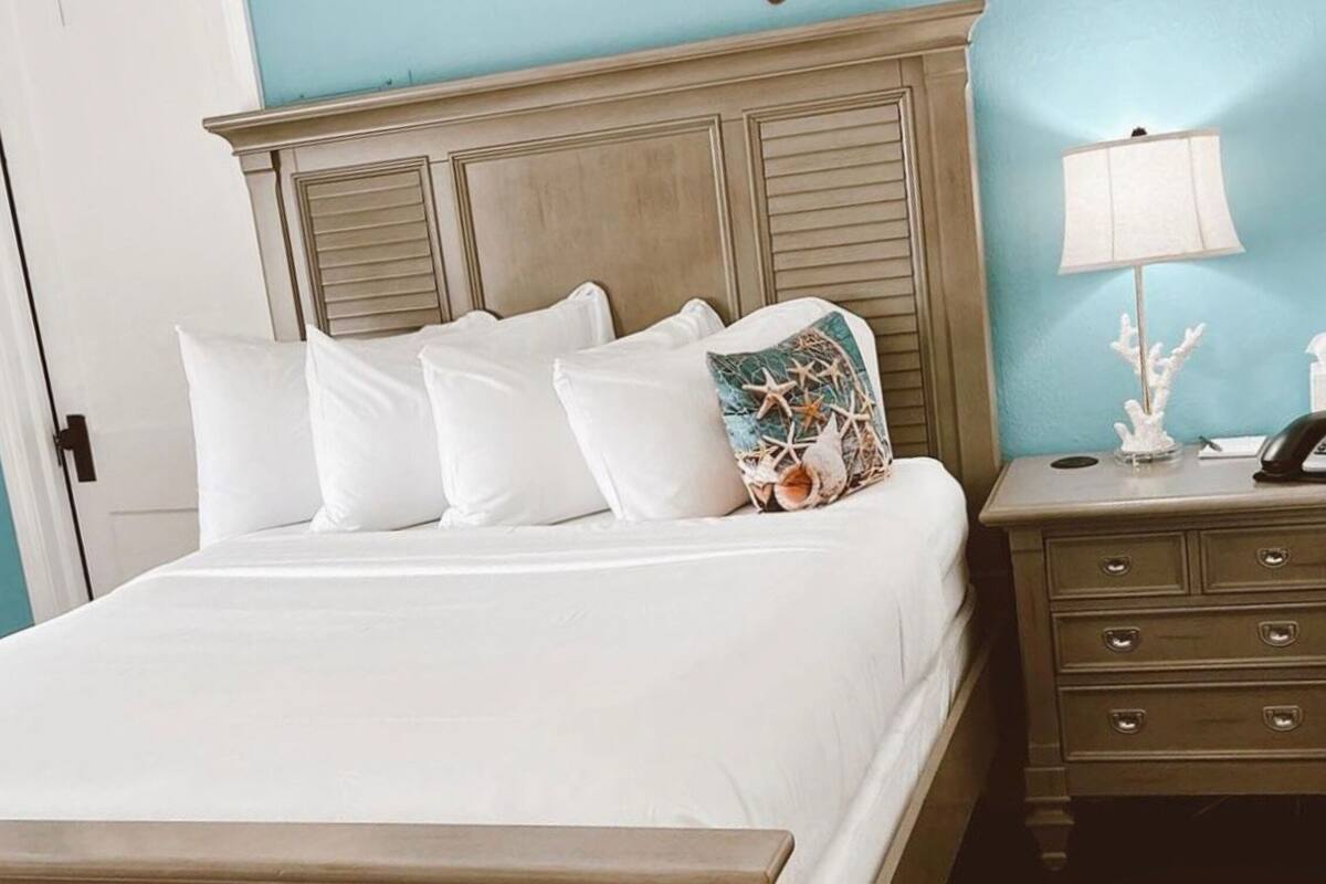 A neatly made bed with white bedding and multiple pillows, a wooden headboard, a bedside table with a lamp, and a decorative coral piece.