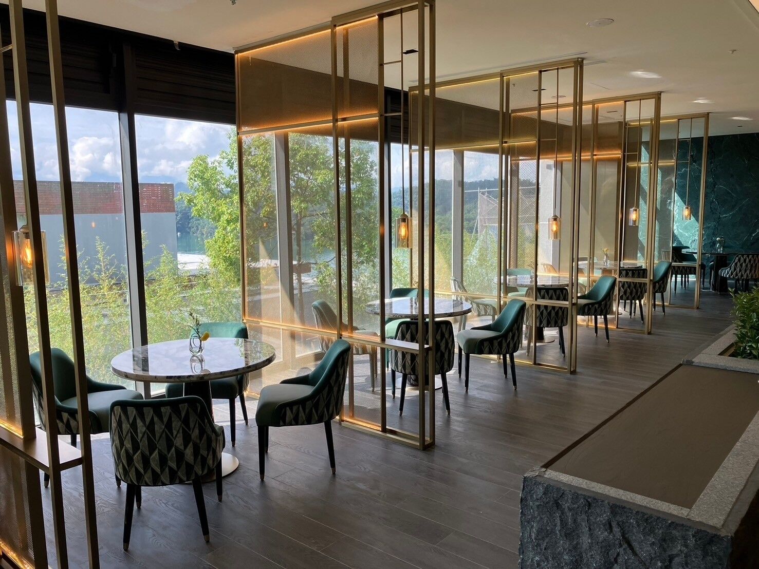 A modern restaurant with round tables and green chairs, large windows offering a view of trees and buildings, and a wooden floor.