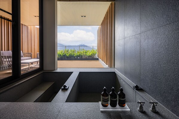 Un baño moderno con una bañera grande, zona de ducha y vistas al mar. Hay tres botellas de productos de baño Aesop en la encimera.