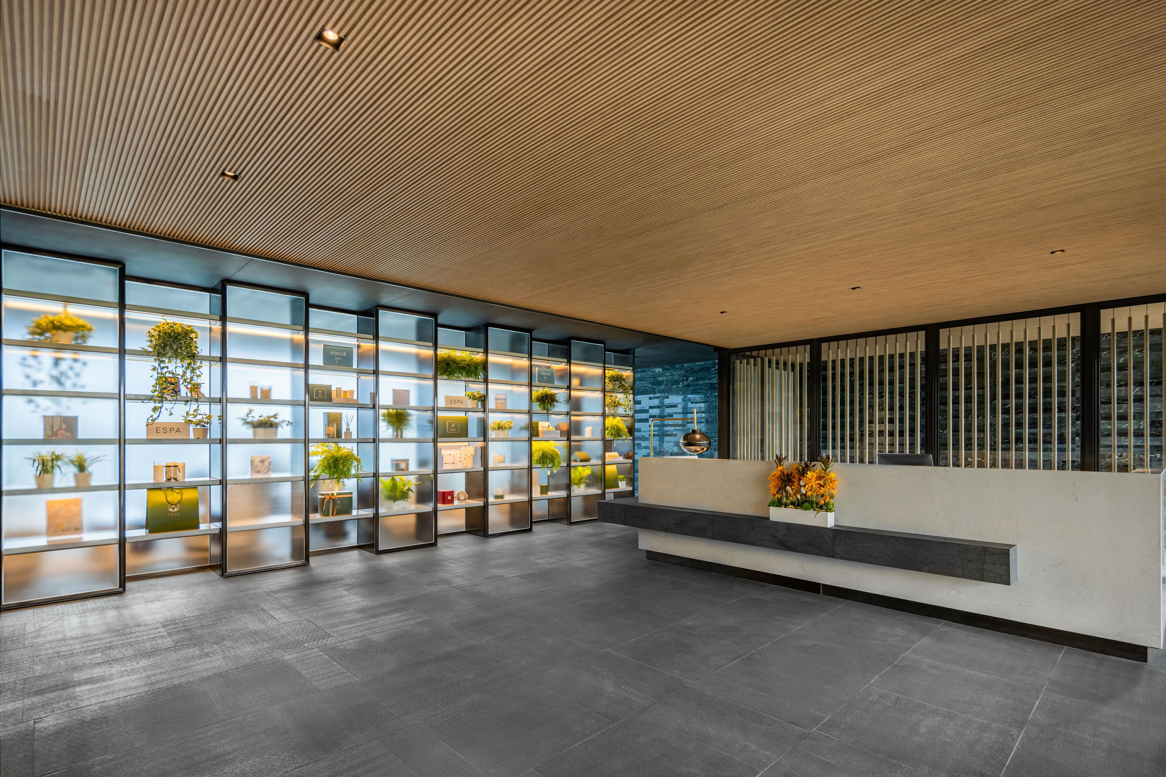 A modern reception area with a wooden ceiling, glass shelving with plants, and a seating area with a vase of flowers.