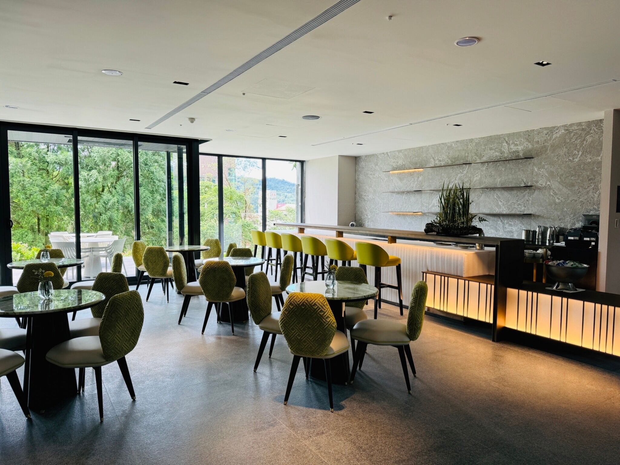 Un restaurante moderno con mesas redondas y sillas amarillas, amplias ventanas que ofrecen vistas a zonas verdes y una zona de cocina con salpicadero de mármol.