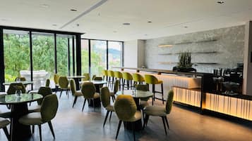 Un restaurante moderno con mesas redondas y sillas amarillas, amplias ventanas que ofrecen vistas a zonas verdes y una zona de cocina con salpicadero de mármol.