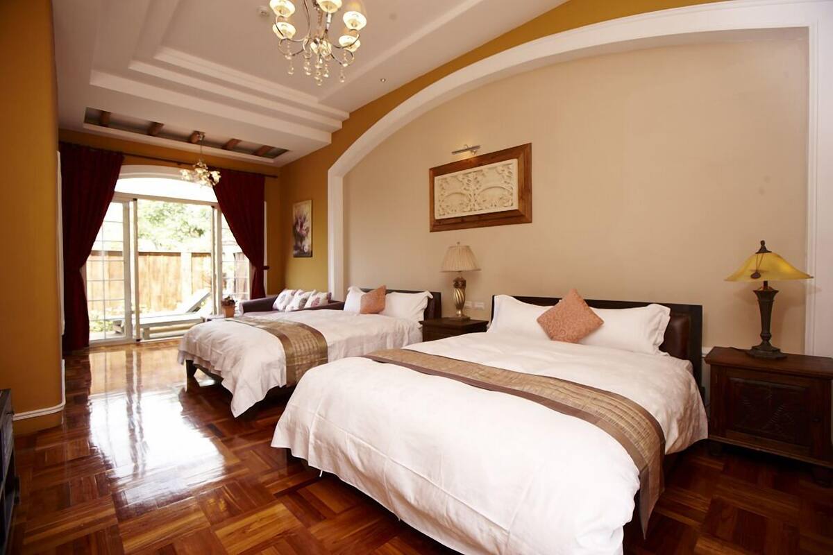 Habitación de hotel con dos camas, piso de madera, un candelabro y un ventanal con cortinas.