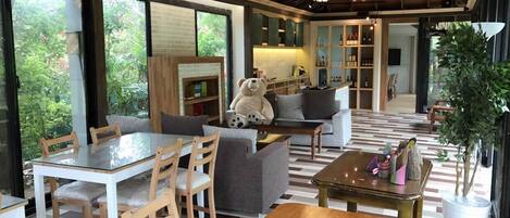 A spacious living area with a dining table, chairs, a sofa, and a large teddy bear. There is a kitchen area with shelves and a refrigerator in the background.