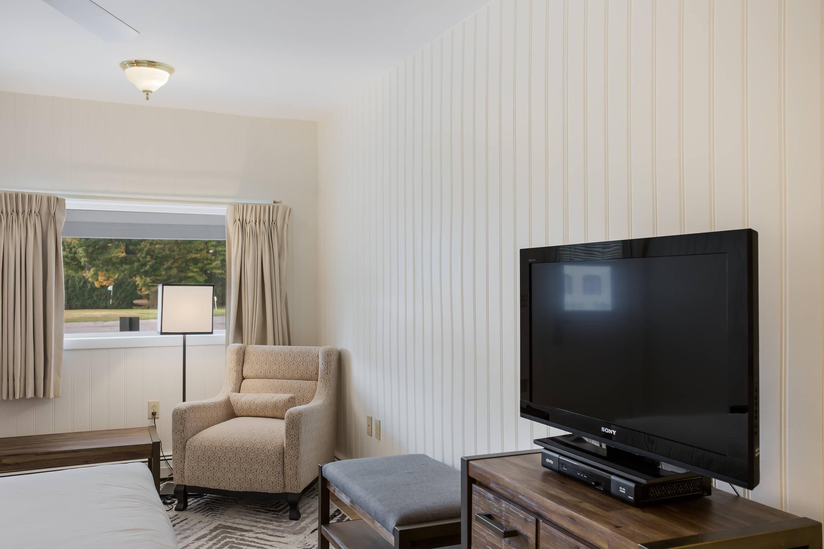 A hotel room with a large flat-screen TV, a comfortable armchair, a bedside table, and a window with curtains.