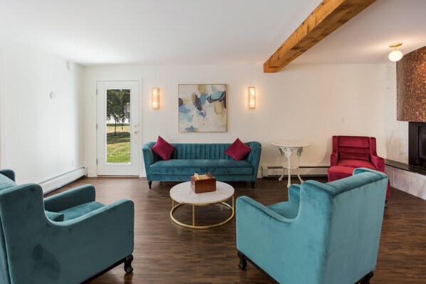 A modern living room with teal sofas, a round coffee table, and a red chair. There is a painting on the wall and a small table with a lamp.