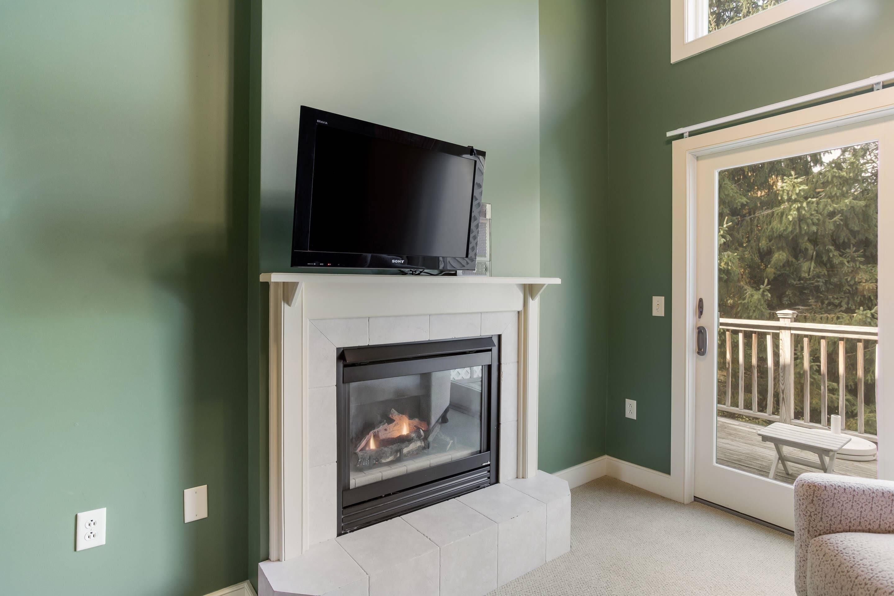 A living room with a fireplace, a television on top, and a view of the outdoors through a sliding glass door.