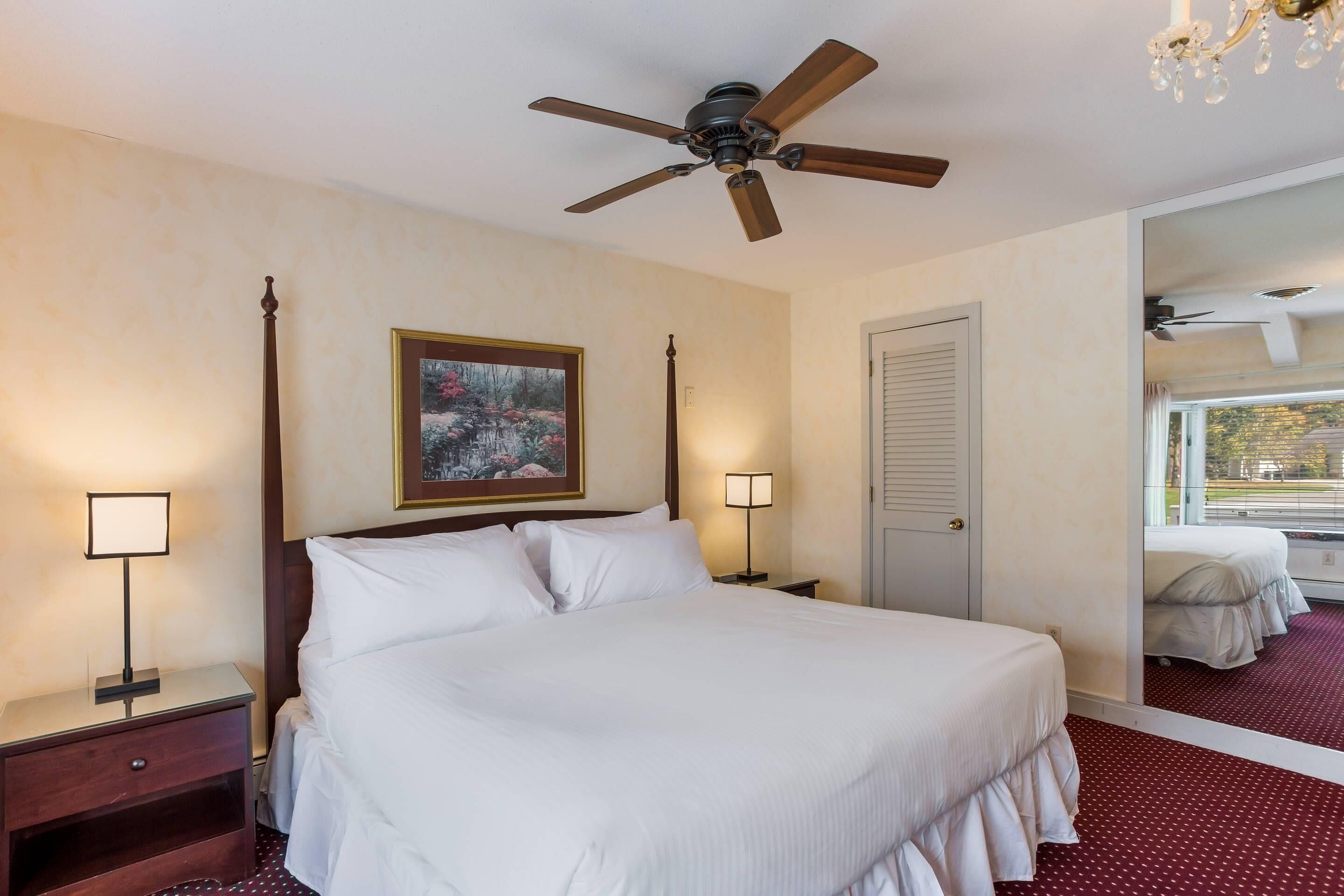 A hotel room with a large bed, a ceiling fan, a nightstand with a lamp, a framed picture on the wall, and a mirror reflecting the room.