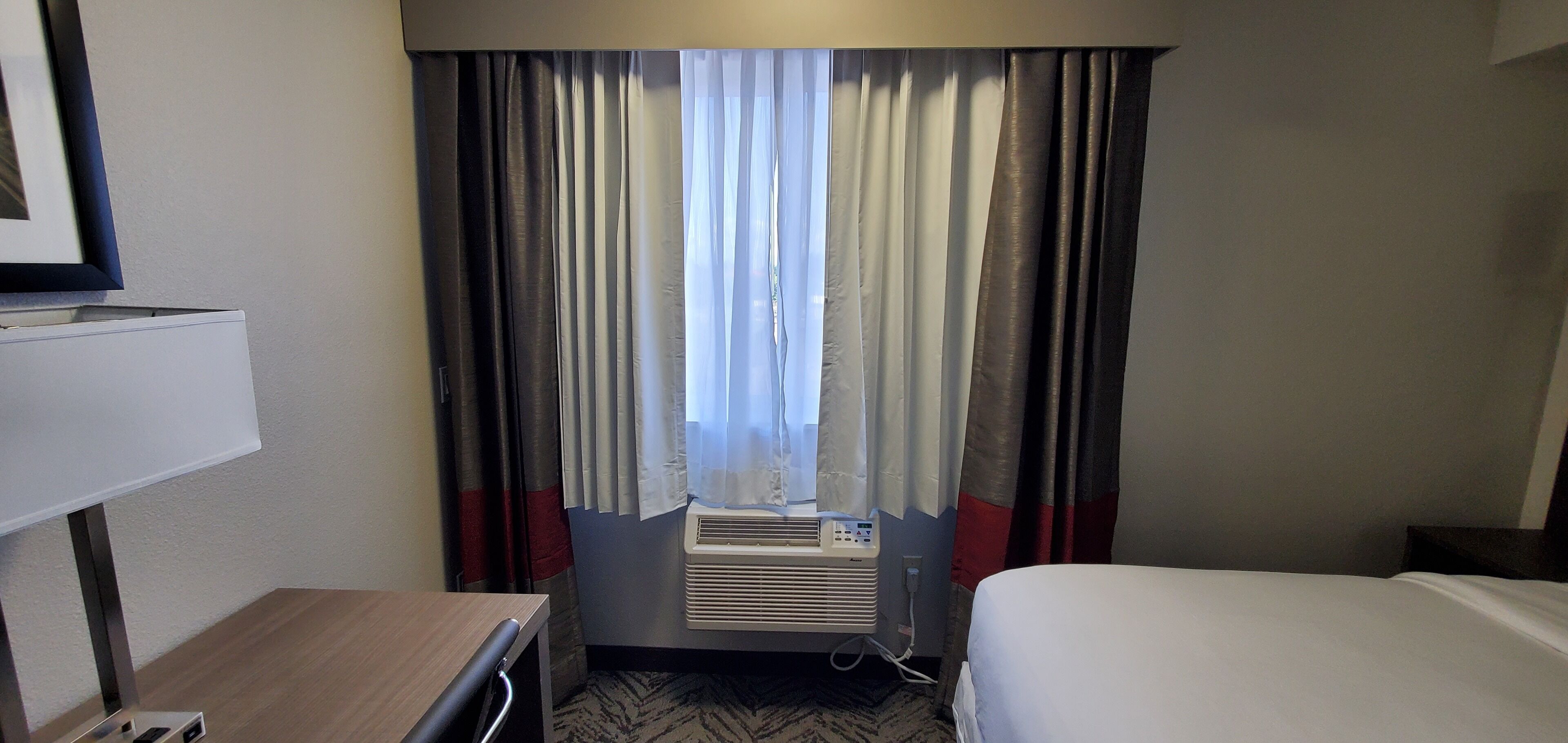 A hotel room with a bed, a desk, a chair, an air conditioning unit, and a window with curtains.