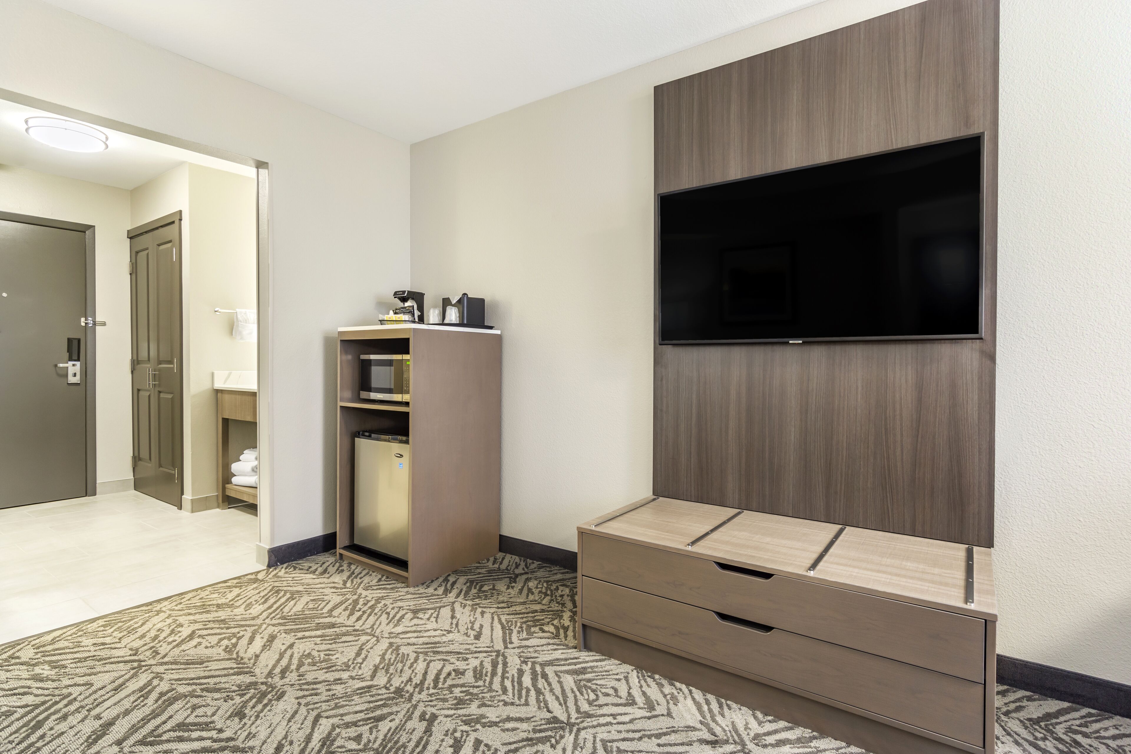 A hotel room with a flat-screen TV mounted on the wall, a wooden cabinet with a microwave, and a patterned carpet.