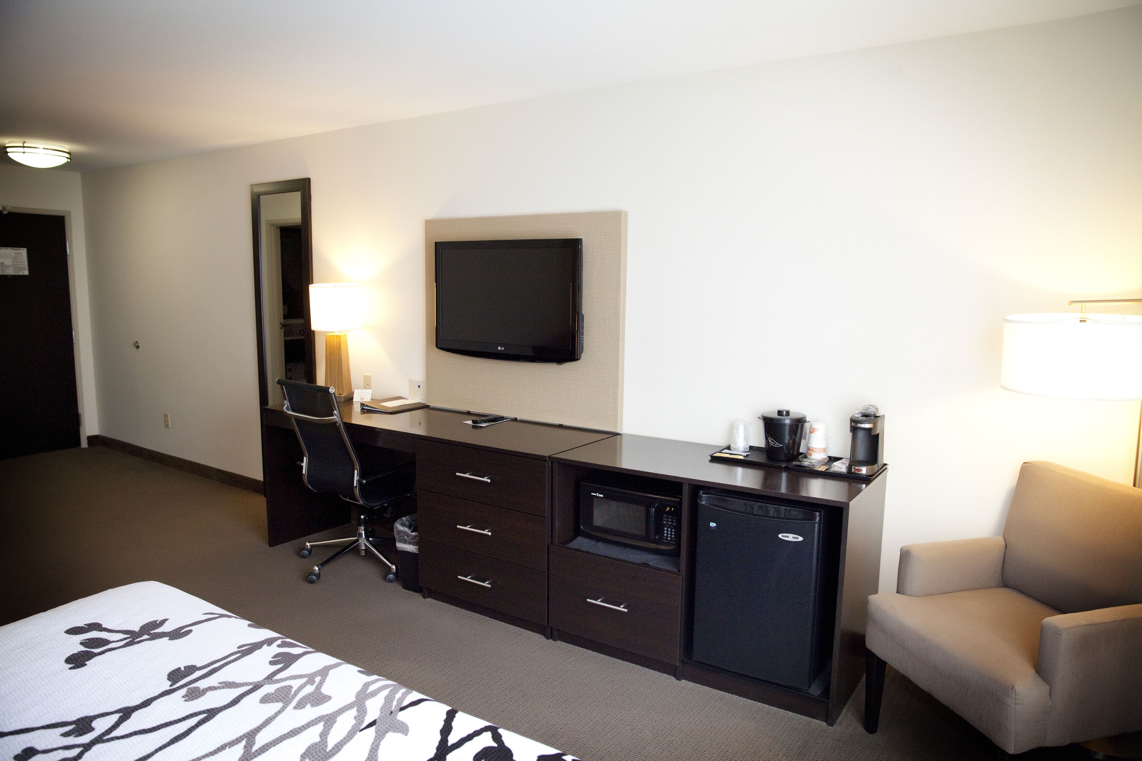 A hotel room with a bed, a desk with a flat-screen TV, a microwave, a coffee maker, a chair, and a lamp.