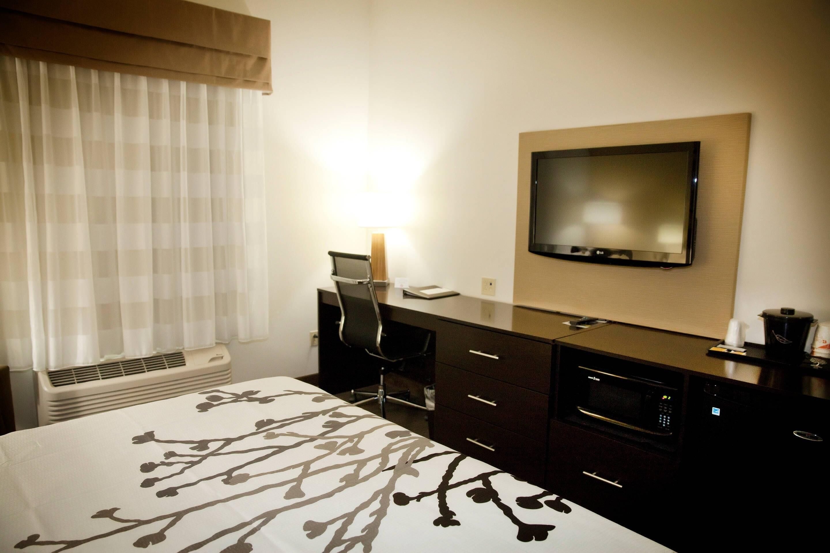 A hotel room with a bed, a desk with a chair, a flat-screen TV, a microwave, and a window with curtains.