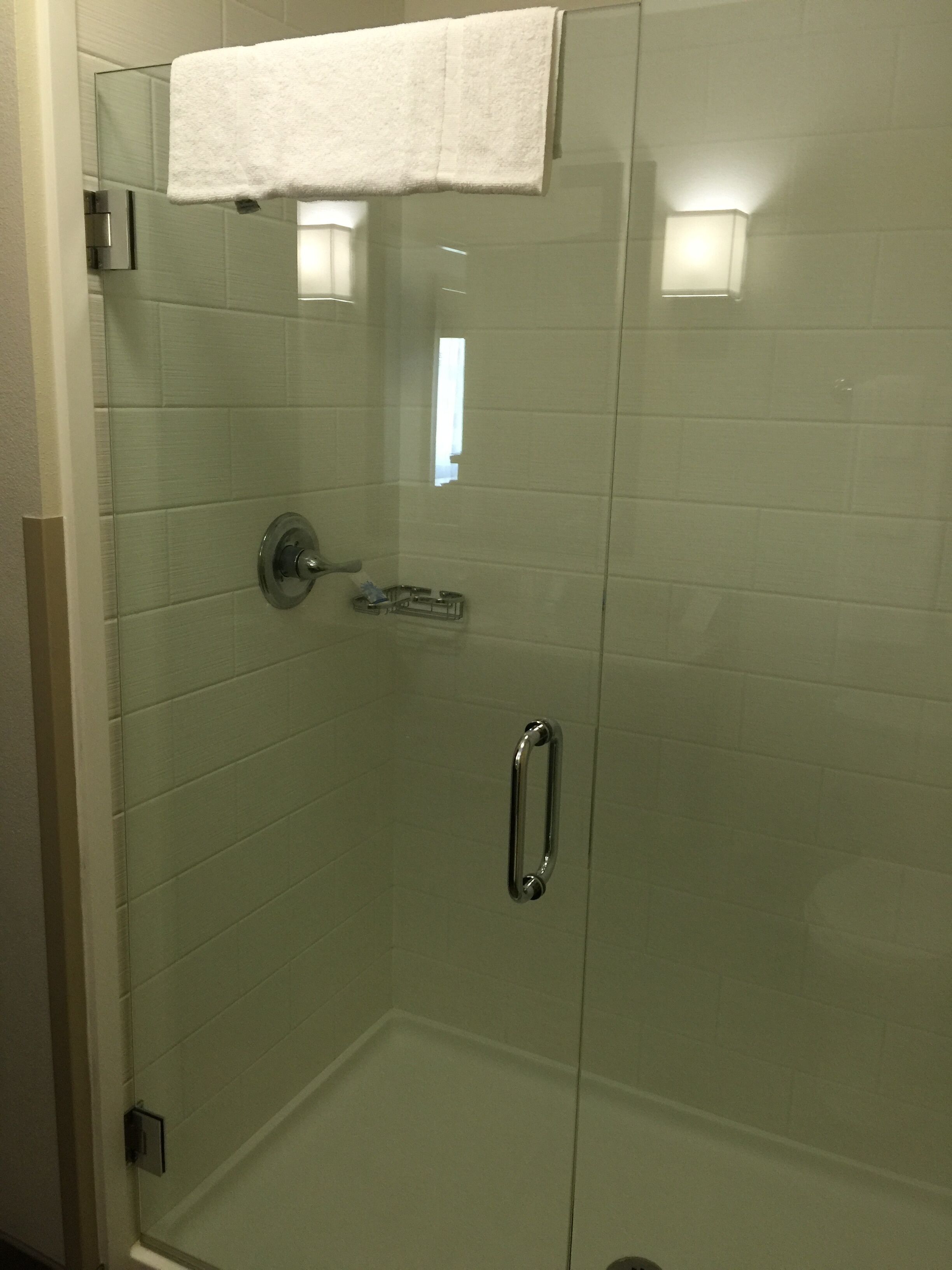 A glass-enclosed shower stall with a white towel hanging on the door, a small window, and a handrail.