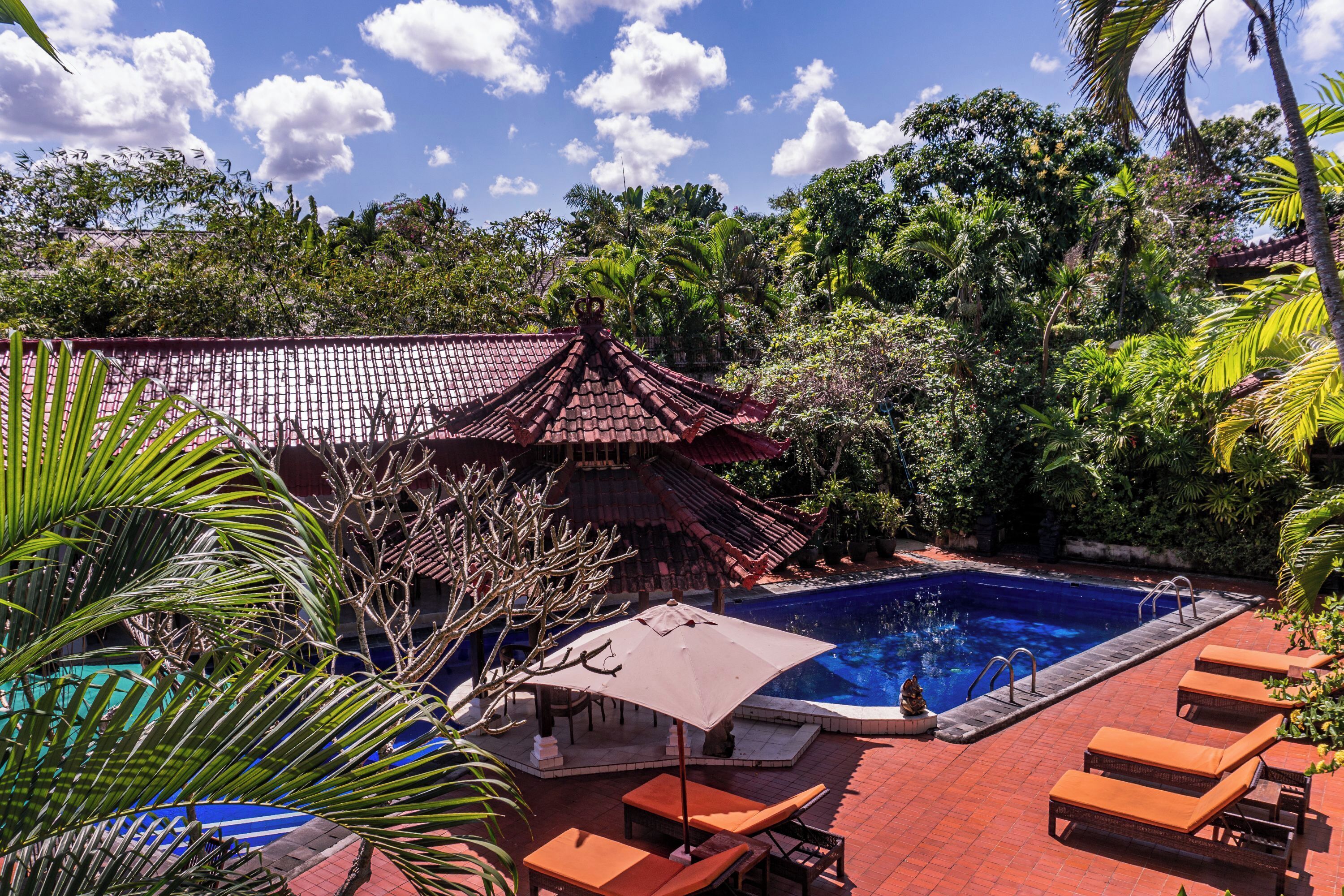 A pool area with lounge chairs and a covered seating area, surrounded by lush greenery and a traditional building.
