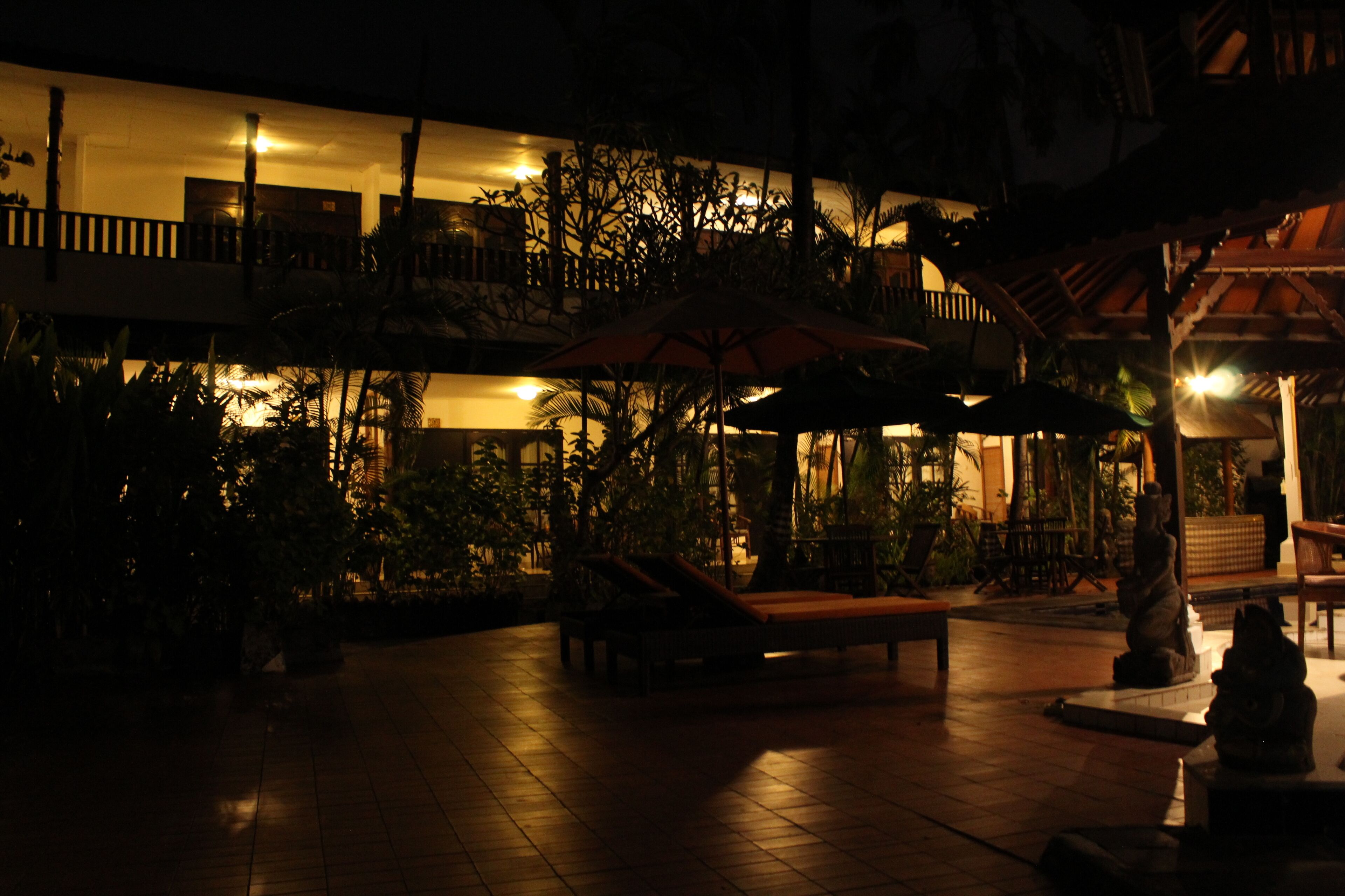 An outdoor area with a tiled floor, lounge chairs, and umbrellas. There are plants and a statue in the foreground.