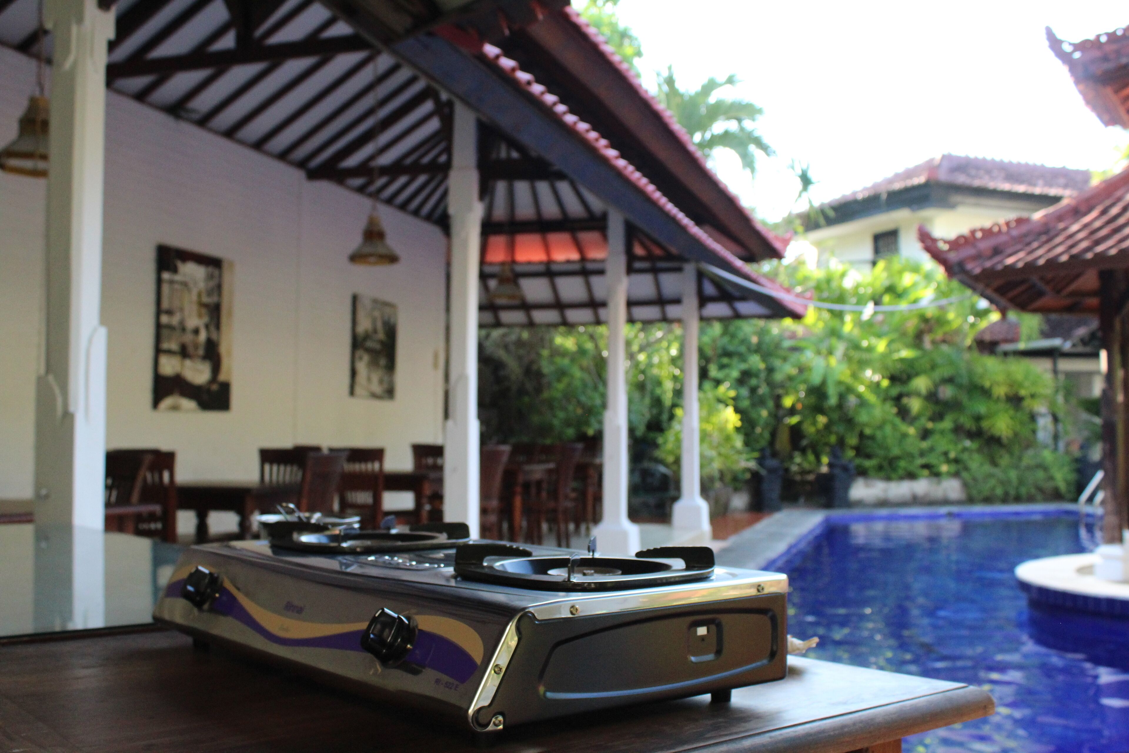 A gas stove on a table near a pool, with a covered outdoor seating area and greenery in the background.