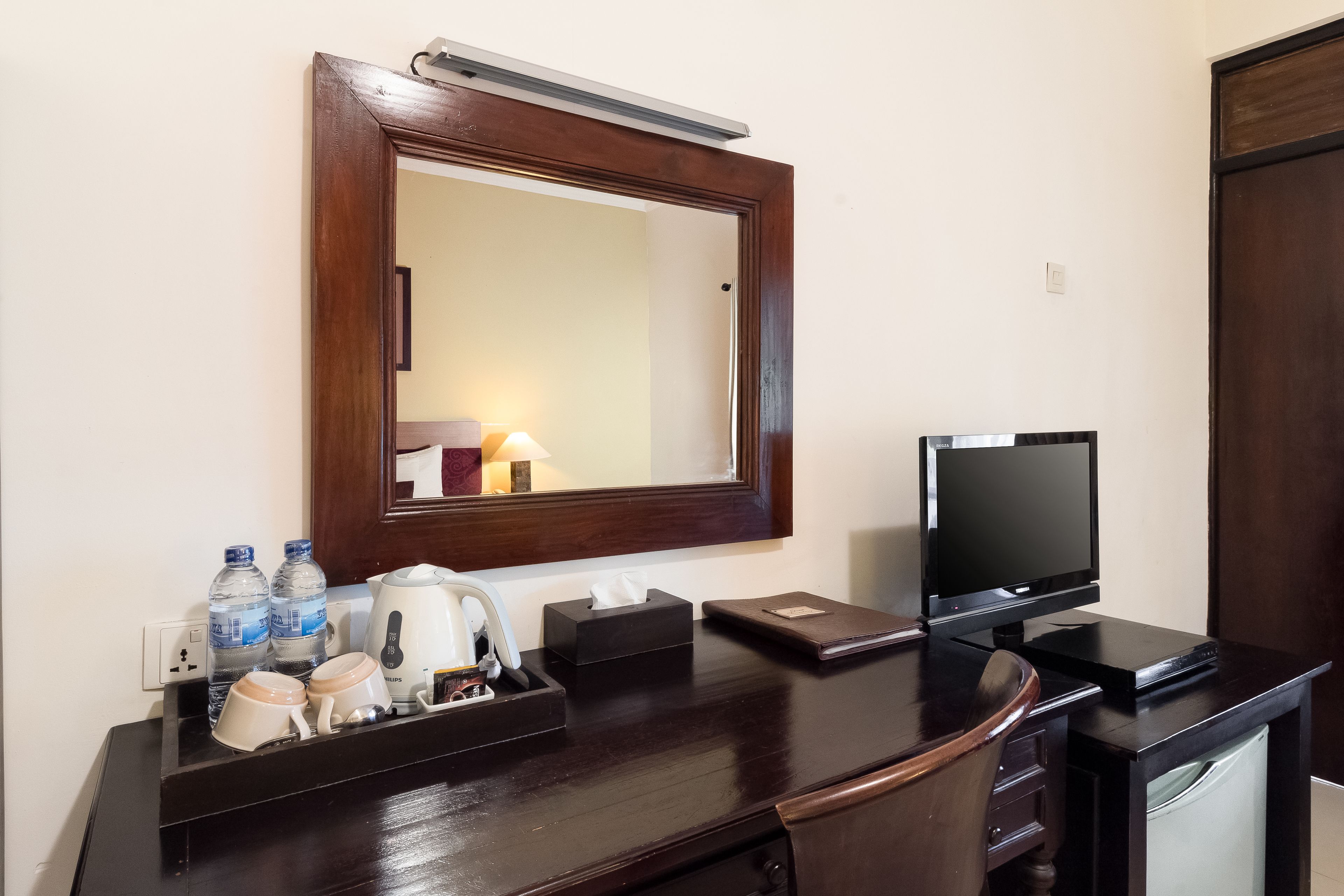 Une chambre d’hôtel équipée d’un bureau en bois, d’un téléviseur à écran plat, d’un miroir, d’une machine à café, d’une boîte de mouchoirs et de deux bouteilles d’eau.